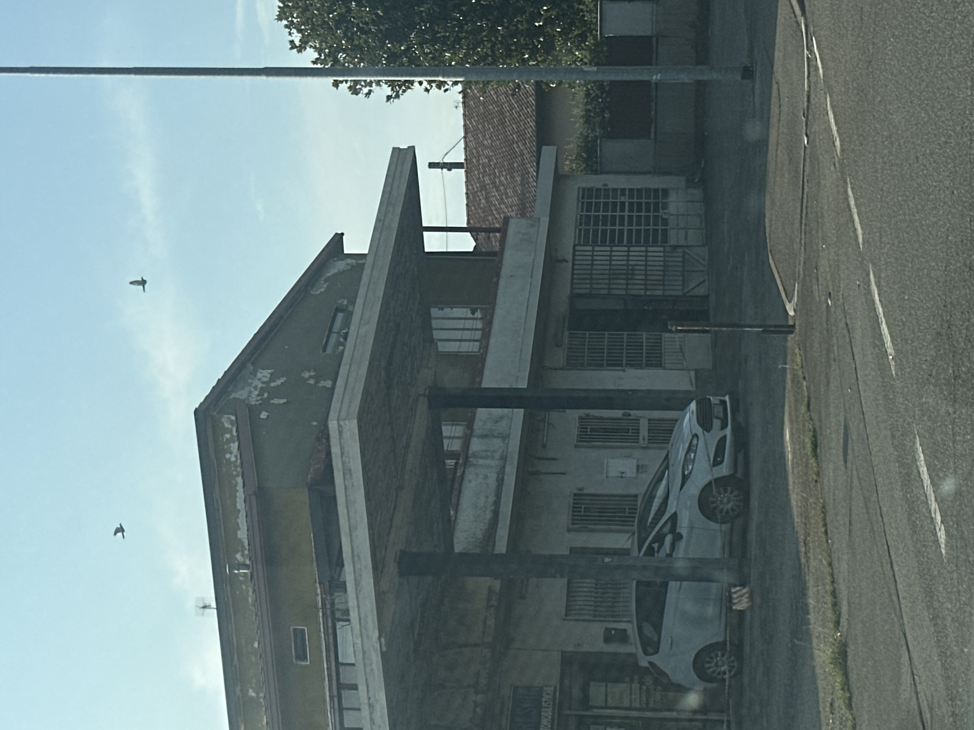 The image shows a street scene in Rudiano, Italy. In the foreground, a grey car is parked under a covered structure. The structure appears to be a covered parking area or part of a building with an overhanging roof supported by pillars. Behind the car, a building with a greenish, weathered facade is visible. The building has windows and doors with metal grates. In the sky, two birds are in flight. A tall, grey utility pole stands to the right of the building, and a tree with dark green foliage is partially visible behind it. The street in front of the covered structure has markings, suggesting it is a road or parking area. The overall environment appears to be an urban or semi-urban setting.