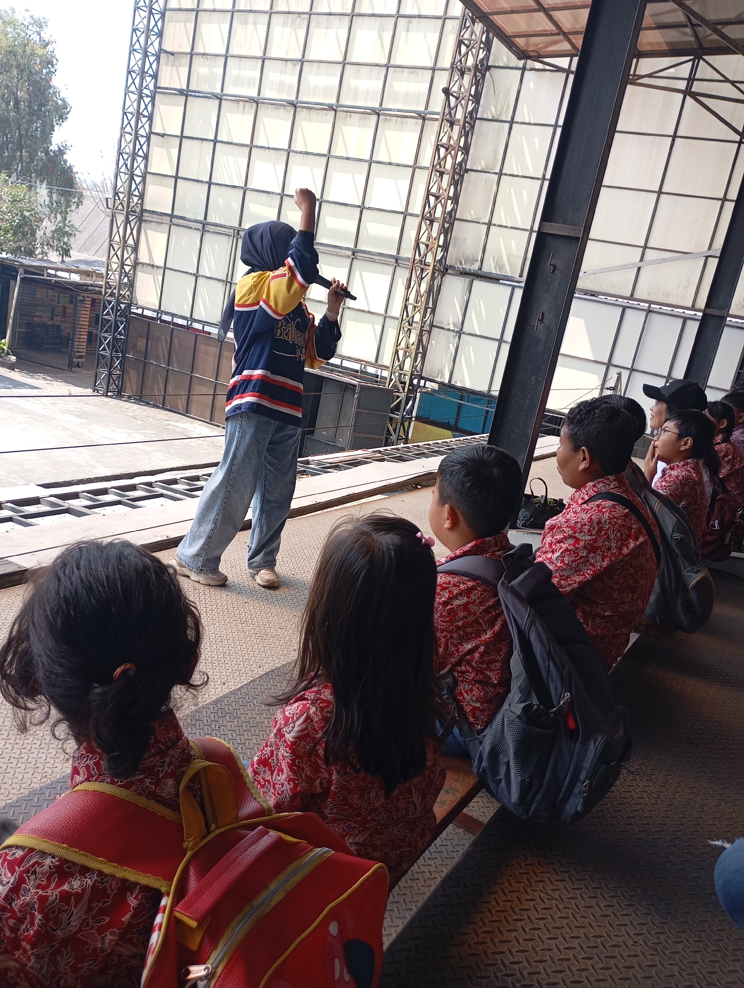 In an outdoor amphitheater setting, likely within the city of Batu, Indonesia, a young woman wearing a hijab, blue and yellow striped long-sleeved shirt, and light-wash jeans stands with a microphone in her right hand, her left arm raised in a fist. She is speaking to an audience seated on tiered benches. The audience consists primarily of children, many of whom are wearing matching red shirts with a white floral pattern. Some of the children have backpacks. The background features a large, open-sided structure with a metal frame and translucent panels, suggesting it is a stage or performance area. The overall scene depicts an outdoor event or educational program.