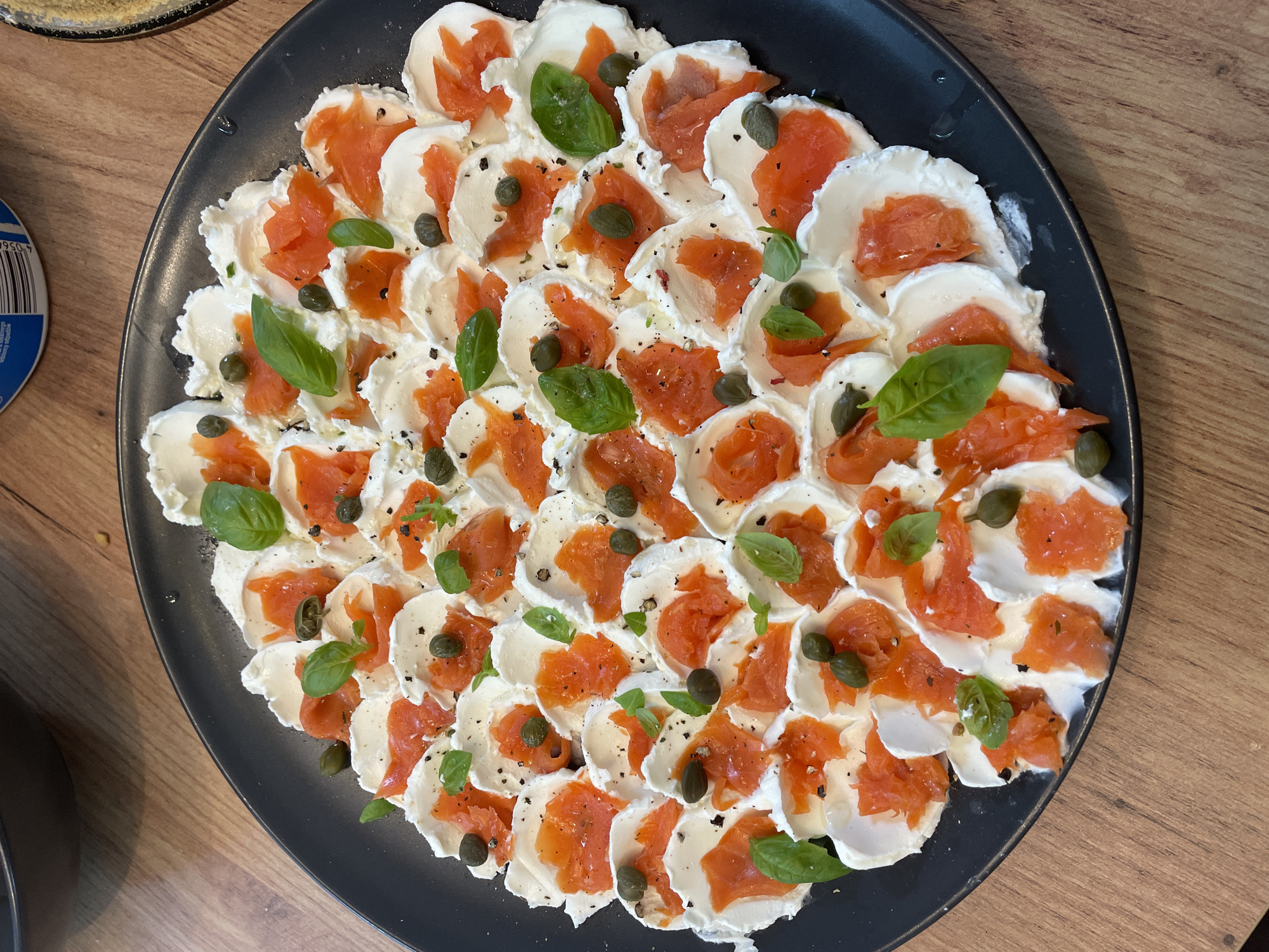 A black ceramic plate filled with a visually appealing appetizer sits on a wooden table. The dish consists of thinly sliced mozzarella cheese arranged in a circular pattern. Each slice of mozzarella is topped with pieces of smoked salmon, a few capers, and a fresh basil leaf. Black pepper is sprinkled across the entire dish. The setting appears to be an indoor environment, possibly a kitchen or dining area, given the wooden table and the suggestion of other objects in the background, though they are out of focus. The lighting suggests an overhead light source.