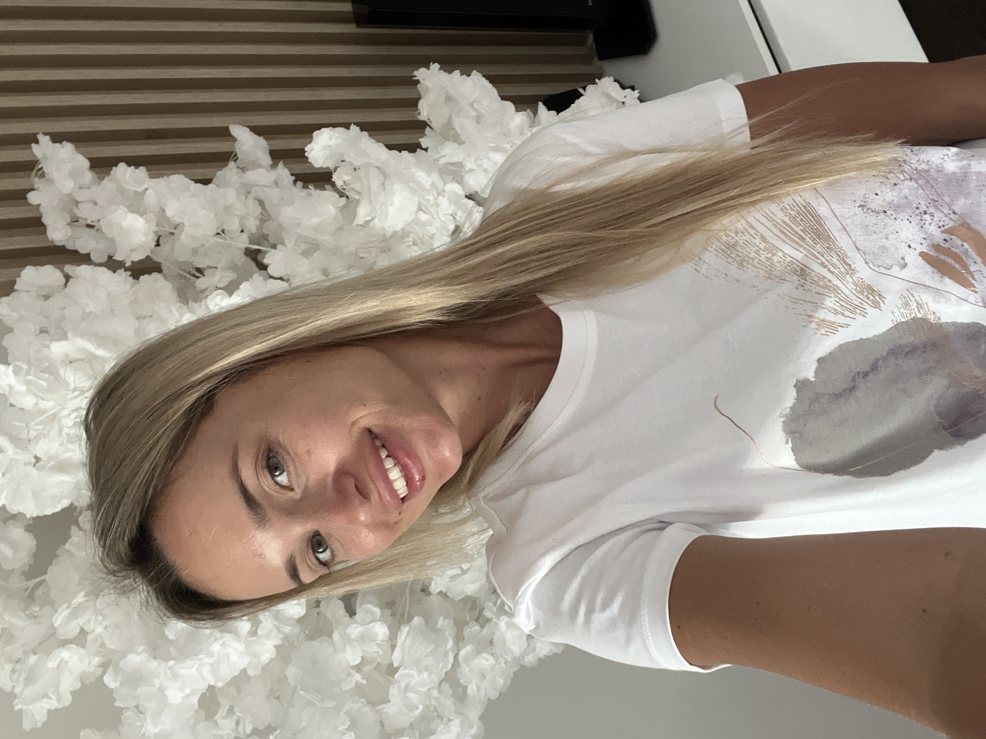 A young, blonde woman with green eyes smiles at the camera. She is wearing a white t-shirt with a light-colored abstract print. She is standing in front of a backdrop of white artificial flowers and a wall with vertical wooden paneling. A piece of black electronic equipment is visible to the right of the woman. The woman's arm is on the left side of the frame. The lighting suggests it is daytime, and the scene appears to be indoors, possibly in a hotel room or a decorated venue in Sharm El Sheikh, Egypt.