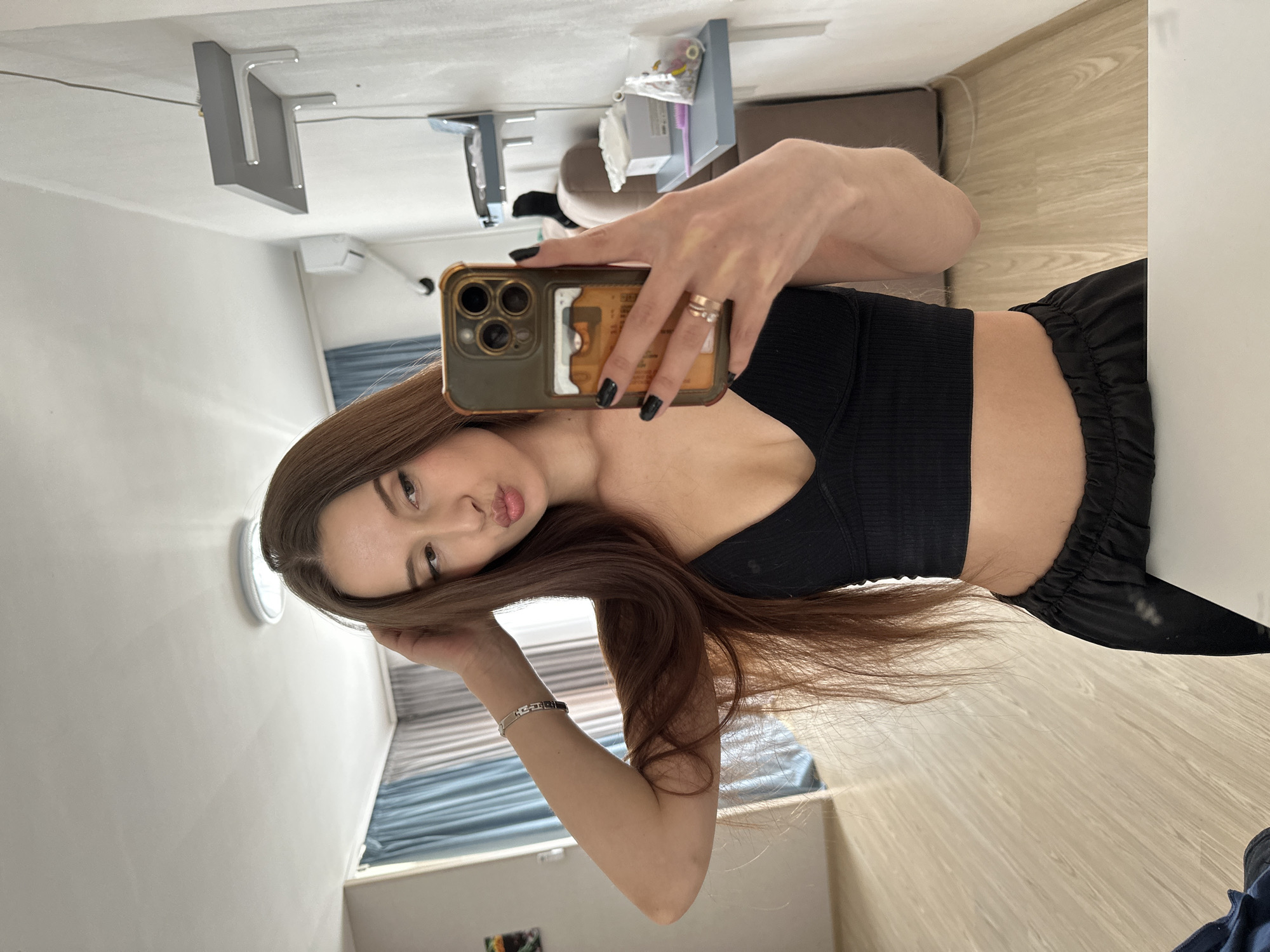 A woman is taking a selfie in an indoor environment. She is wearing a black crop top and dark bottoms. She is holding an iPhone, likely to capture the image. The background shows a room with light-colored walls and wooden floors. In the periphery, a bed with blue-grey linens is visible. The room appears to be located in an apartment or residence within Yekaterinburg, Russia, as this is the specified location.