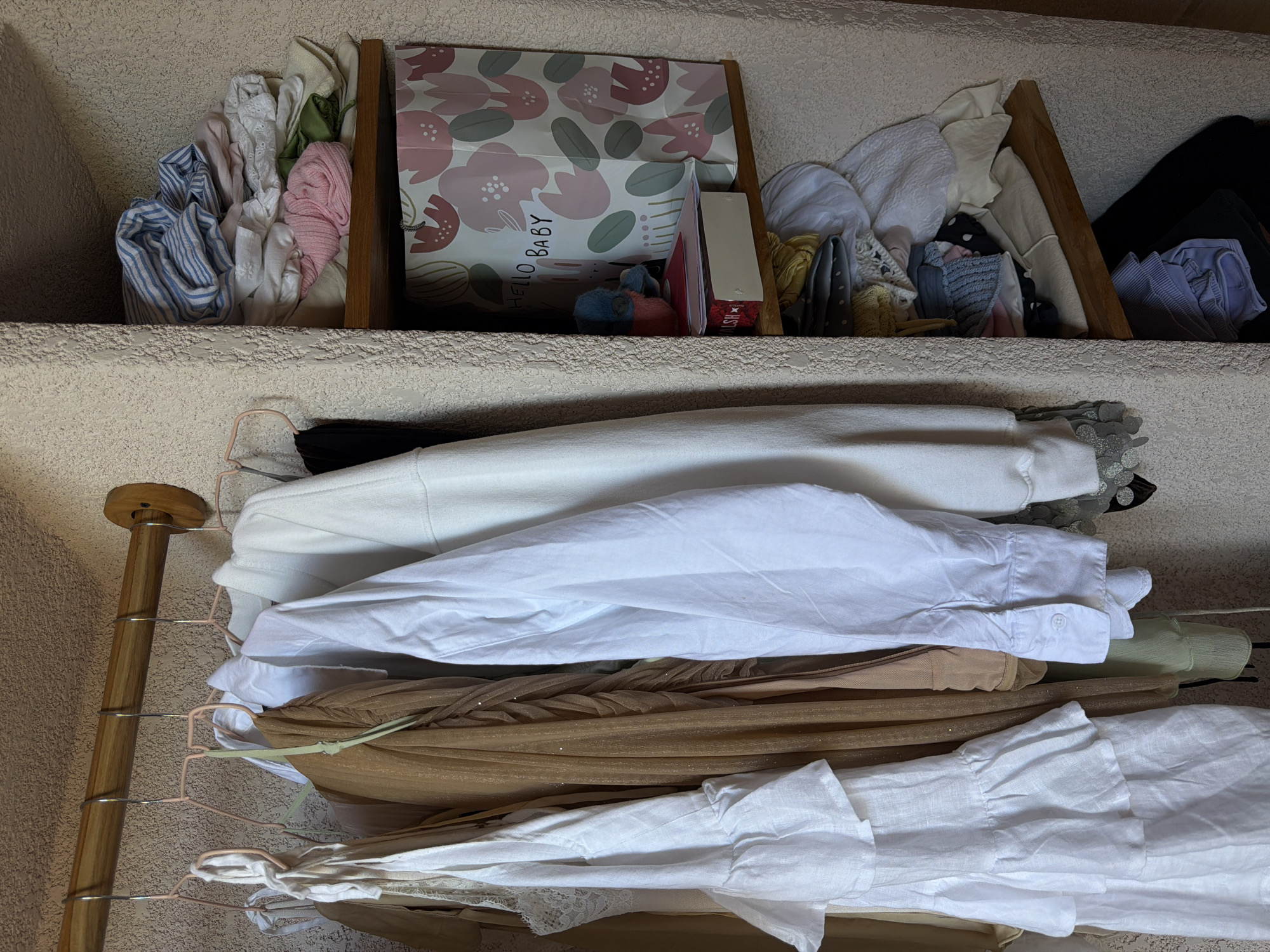 A close-up, eye-level shot showcases a section of a closet with clothing hung on a wooden rod and stacked on wooden shelves. On the left, a white linen dress with ruffled layers is visible, along with other garments in muted tones. To the right, shelves are filled with folded clothes, including a stack of blue and white striped fabric, a pink knitted item, and a gift bag that reads "HELLO BABY" with a floral pattern. The background is a textured wall. This image is located in the city of Mengwi, Indonesia.