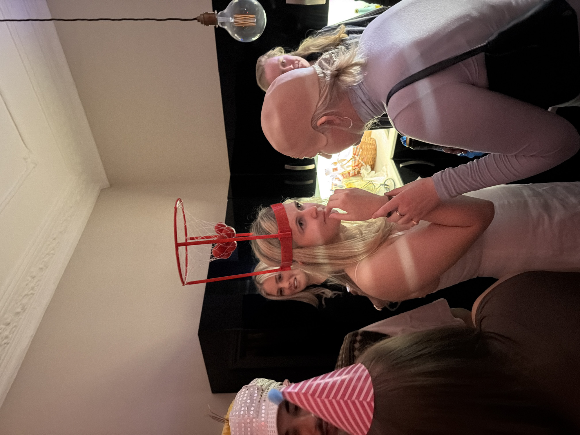 The image captures an indoor gathering of individuals, possibly a birthday celebration given the presence of a party hat and a miniature basketball hoop attached to a person's head. The setting appears to be a domestic interior with white walls and decorative molding. A hanging light fixture with an exposed bulb is visible. At least five people are identifiable, including two women with blonde hair who seem to be the focus of the image. They are positioned close to one another and engaging in a conversation. A third person can be partially seen wearing a party hat. The image does not reveal any identifiable features indicating that this image was taken in London. It is therefore impossible to identify any landmark in London.