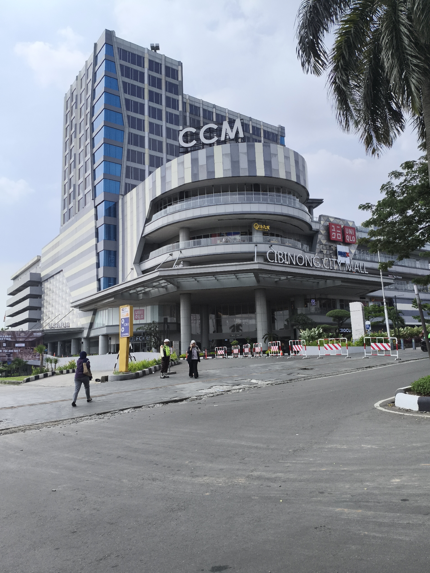 This outdoor daytime image showcases the modern exterior of Cibinong City Mall in Cibinong, Indonesia, under an overcast sky. The complex comprises a tall, multi-story building featuring the letters "CCM" at its peak, and an attached, curvilinear mall structure with "CIBINONG CITY MALL" prominently displayed on its facade. The architecture incorporates extensive use of glass and striped paneling in shades of grey and blue.
Several recognizable store logos are visible on the mall, including "UNIQLO" and "MAX," along with the characters "清品屋" and "Tea House." To the left, a large banner partially reveals information about "HARRIS" (likely a hotel) and an "ALL DAY MEETING" promotion for "R 700.000 NET."
In the foreground, an asphalt road curves towards the mall entrance. Red and white striped traffic barriers line the edge of the pavement, suggesting controlled access. Three individuals are present: one person walks away from the camera on the pavement, while two others, possibly mall staff or security personnel (one in a high-visibility vest), stand near a yellow directional sign. This sign indicates "CCM2" and a "LOADING" area, also featuring the "UNIQLO" logo. Lush green trees and palm fronds frame the scene, adding a natural touch to the commercial setting. The scene depicts a typical commercial environment with ongoing activity.