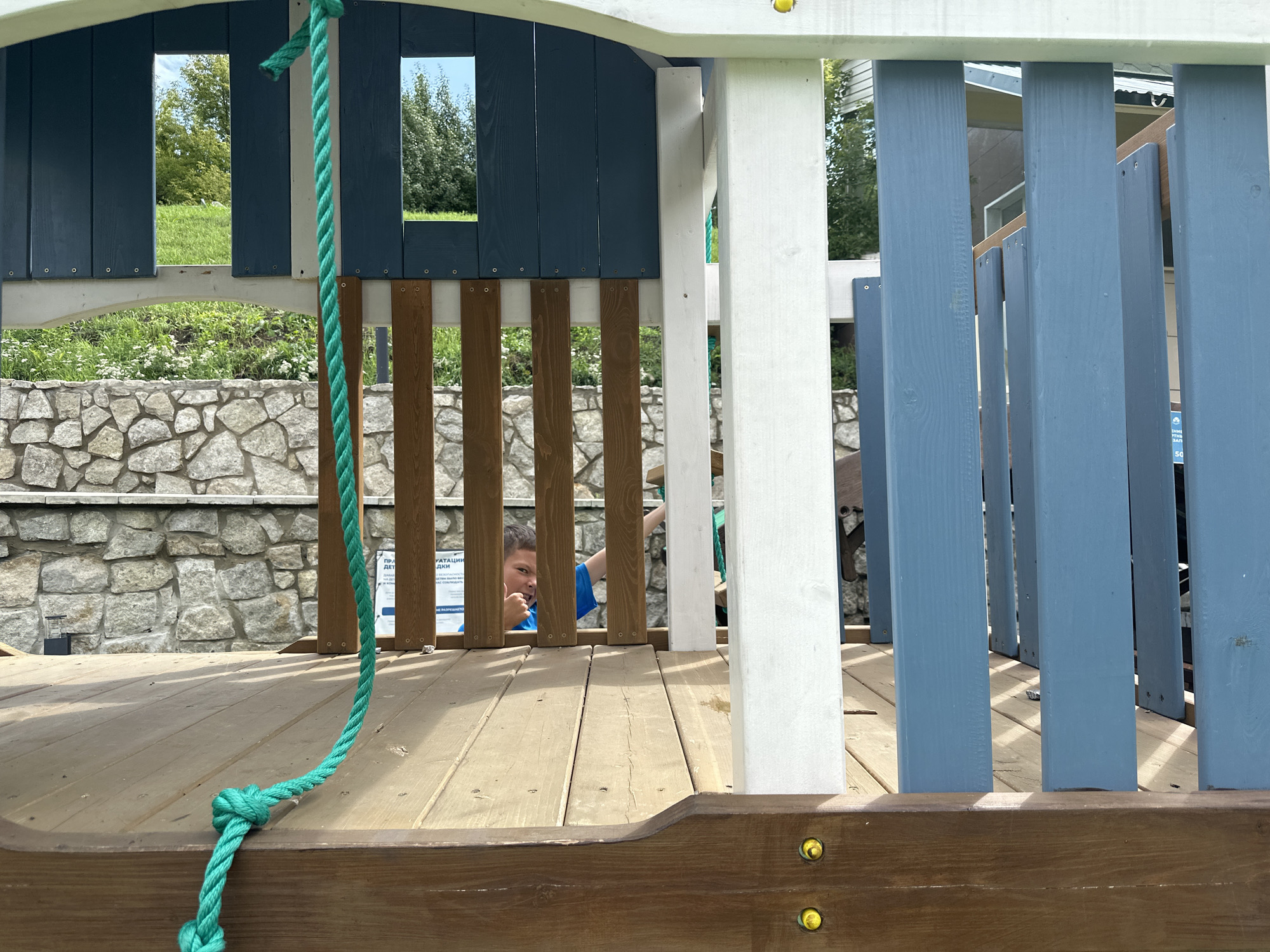 A young boy with brown hair is peeking out from behind the wooden planks of a playground structure. He is wearing a blue t-shirt and giving a thumbs-up gesture. The playground is made of wood and painted in blue and white. A green rope hangs down from the structure, tied to the wooden floorboards. In the background, a stone wall and lush green trees are visible under a clear sky. The setting appears to be a residential area in Danilovka, Altai Krai, Russia.