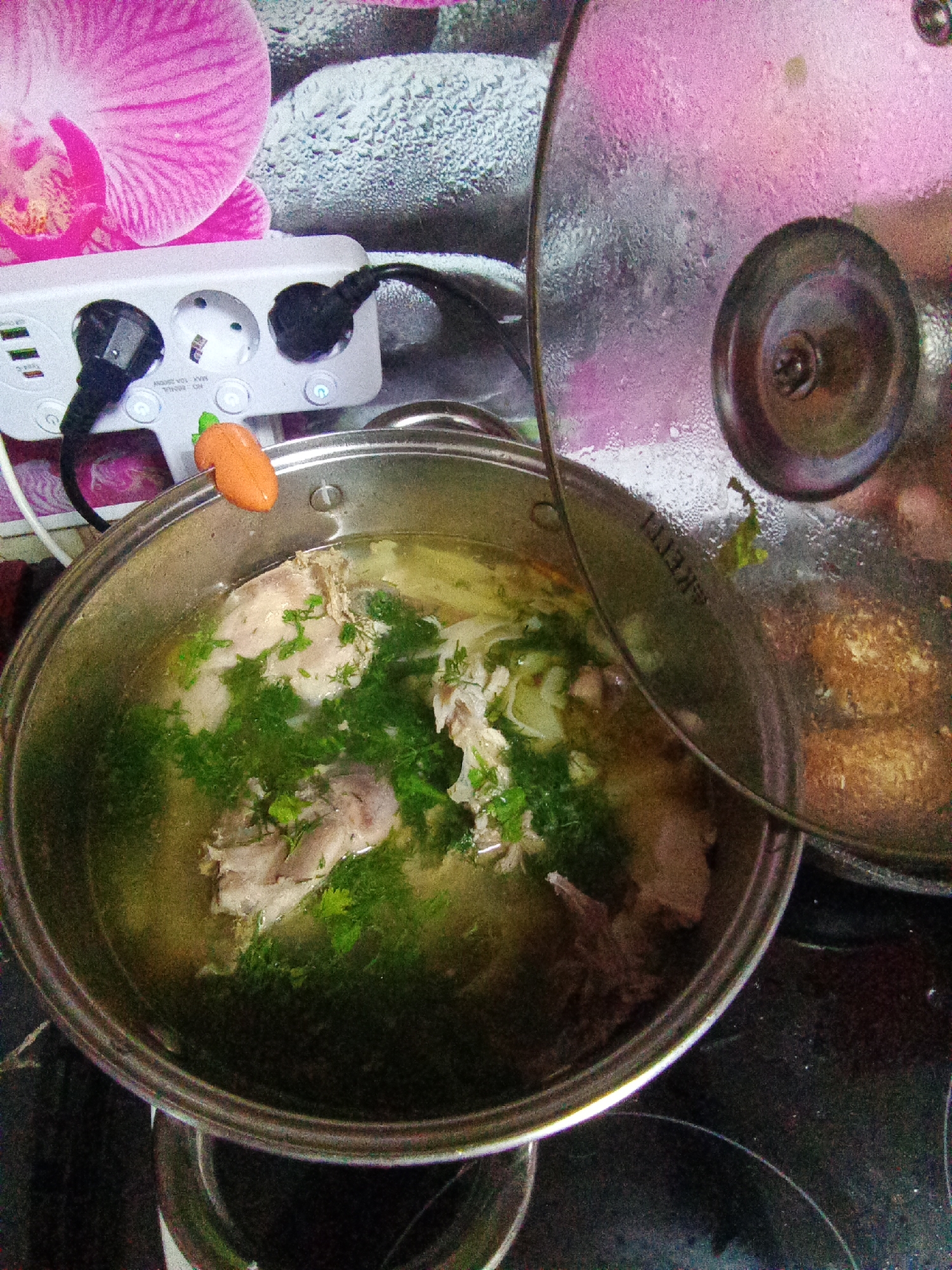 The image depicts a close-up, overhead view of a pot of soup with pieces of meat and green herbs in it, situated on a stovetop. A glass lid, partially open, reveals steam rising from the soup. In the background, there is a power strip with several outlets and a decorative element resembling a carrot clipped to the rim of the pot. The wall behind the power strip features a floral pattern. The overall scene suggests a domestic kitchen environment. The image does not contain any identifiable landmarks or context that would allow for a specific location in Belogorsk, Russia, to be determined.
