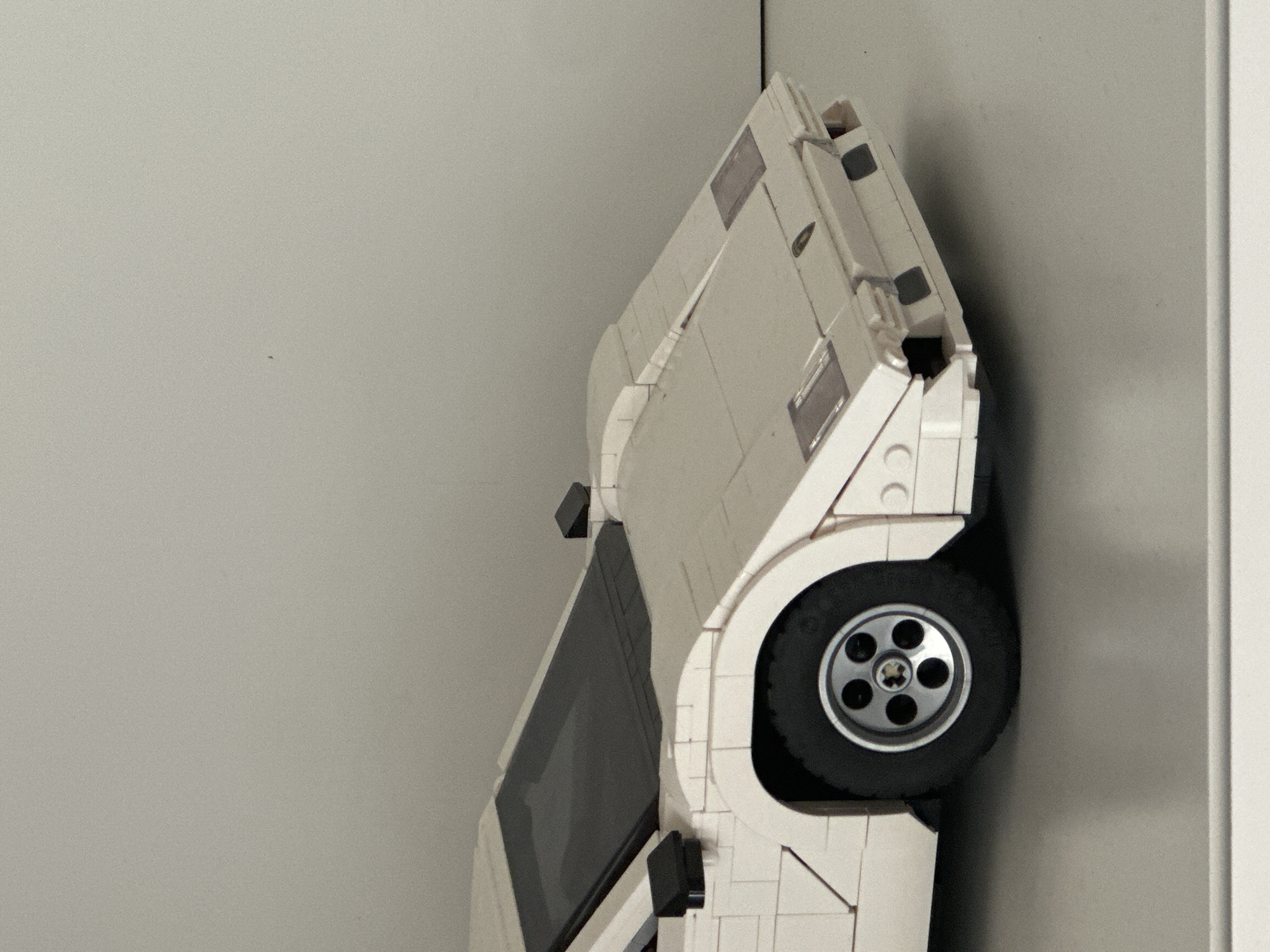 The image captures a white, brick-built model car, likely constructed from LEGO or a similar system, positioned vertically on its side. The car's design suggests a vintage or rally vehicle, featuring a prominent wheel with a multi-spoke rim and a black tire on the right side of the frame. Black rectangular elements are visible on the car's body, possibly representing windows or other details.
The model is leaning against a light-colored vertical surface, which appears to be a wall or a corner on the right side of the image. The rest of the background is a plain, light gray, suggesting an indoor setting. The lighting is even and diffused, without strong shadows, indicating an artificial light source or soft ambient light.
There are no people present, and no active event is taking place; the car is statically displayed. No visible text is present on the model car or in the background. While the provided context indicates the location is Milan, Italy, the image itself contains no specific visual cues to confirm this.