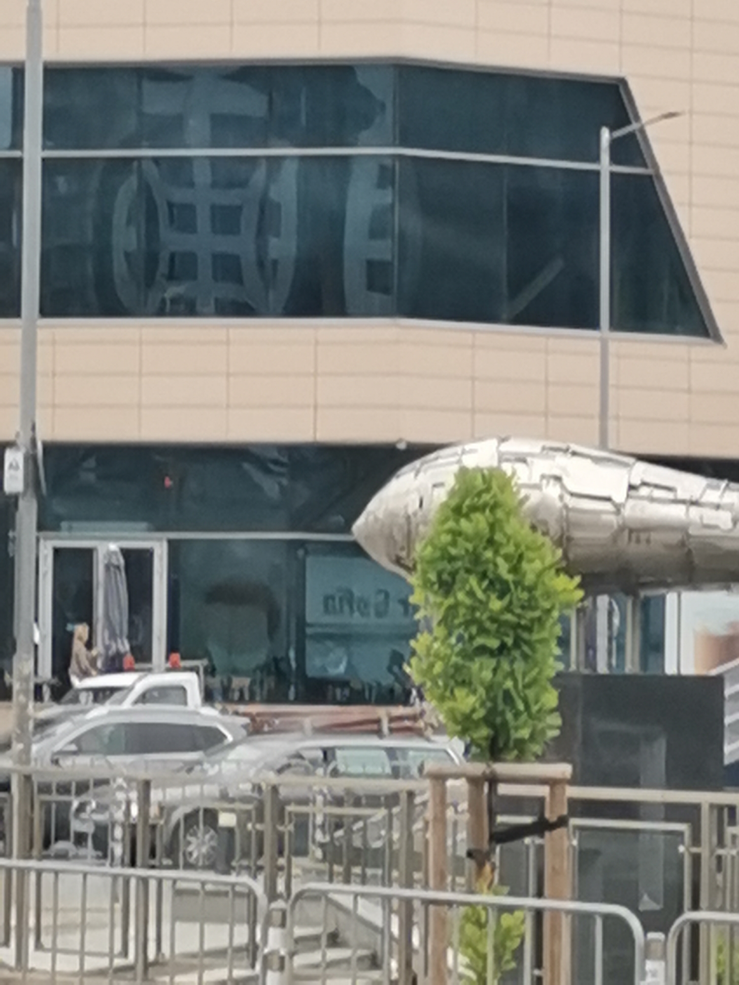 The image shows a metallic sculpture resembling a fish or submarine, positioned in front of a modern building with large, reflective windows. A small, leafy green tree stands in front of the sculpture. In the background, several cars are visible, and some outdoor seating areas with umbrellas are set up in front of a storefront. The building appears to be an office or commercial complex. The reflection in the windows shows a geometric pattern. The scene suggests an urban setting in Sofia, Bulgaria, likely a public space or commercial area.