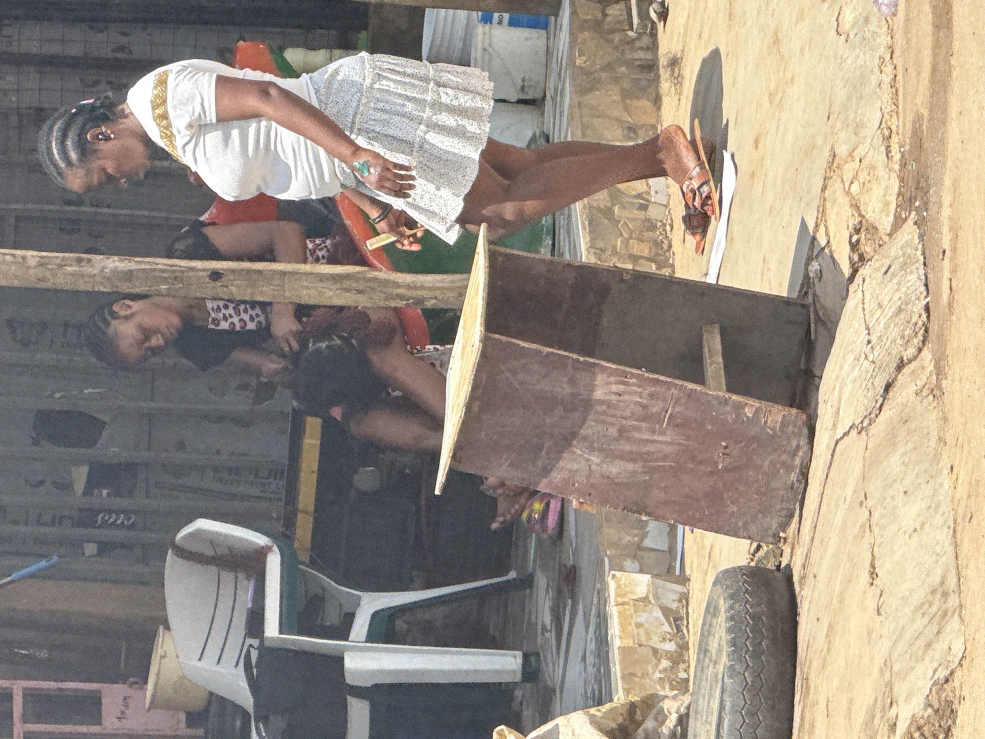 In Abuja, Nigeria, two women are in an outdoor setting with a dirt and concrete ground. The woman on the right, wearing a white t-shirt with gold trim and a patterned skirt, stands with her right foot resting on a flat, dark brown object. Her left foot is bare. She holds a small yellow object, possibly a comb, in her right hand and has a green tattoo on her left hand. The woman on the left, wearing a black jacket over a patterned dress, is seated and appears to be engaged in some activity, possibly braiding hair, with her head bowed. In the foreground, a dark, circular object, resembling a tire, is visible on the cracked concrete ground. Behind the women, there are several plastic buckets, chairs, and a wooden structure. The background shows a building with a textured wall.