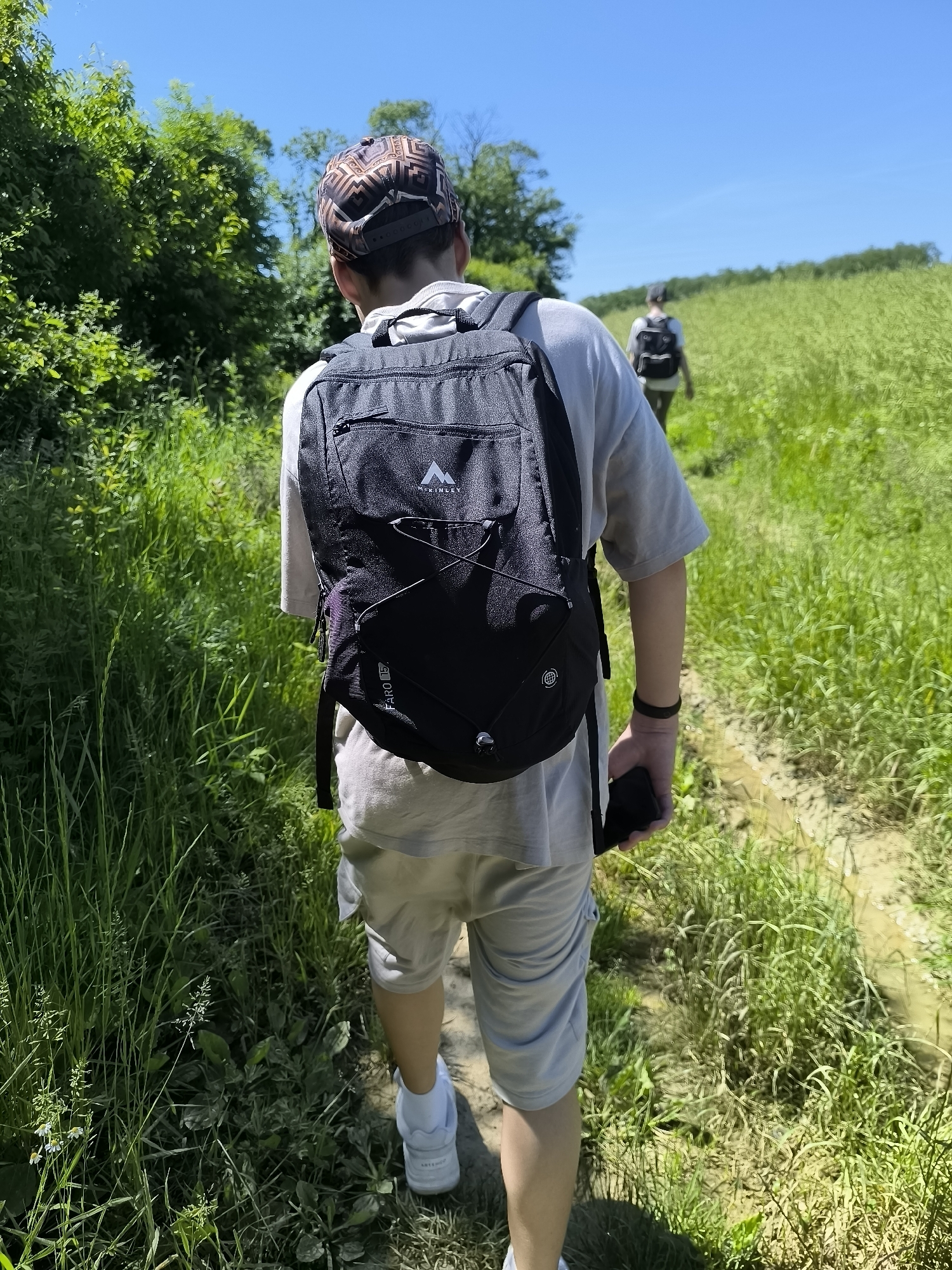 The image depicts two individuals walking along a narrow, muddy path flanked by tall green grass and vegetation under a clear, bright sky. The foreground figure, seen from the rear, wears a patterned cap, a light gray t-shirt, light gray shorts, and white sneakers. They are carrying a black backpack and holding a black object, possibly a phone, in their right hand. A black wristband is visible on their left wrist. The second individual is further down the path, also wearing a backpack. The location is likely Chaponnay, France, suggested by the type of vegetation and rural landscape. The scene suggests a hike or walk in a rural or semi-rural setting.