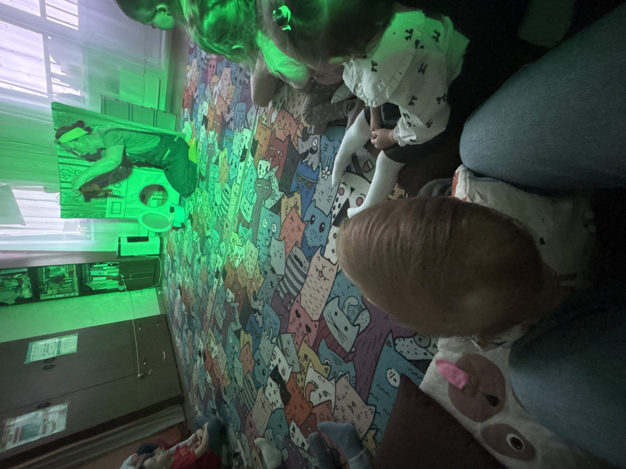 The image captures an indoor scene, likely a playroom or living room, intensely illuminated by a green light source during the daytime, with natural light visible through a window on the left. The floor is covered with a vibrant, colorful rug adorned with numerous cartoon cats in various poses and expressions.
Several people, including children and at least one adult, are present in the room. In the foreground, parts of individuals are visible, including a person with light brown hair wearing jeans, and a child dressed in a white patterned top and white tights. Another child in a red shirt and blue socks is lying relaxed on the cat-patterned rug further back.
On the left side of the room, bathed in the green light, a person wearing a dark shirt, pants, and a headband is leaning over a play structure, possibly a cardboard play kitchen or an appliance mimic. This individual appears focused, engaging with a circular component within the structure. Other heads, illuminated by the green glow, suggest more people are gathered, possibly observing the activity.
A white electronic device, resembling a projector or a specialized lamp, is visible on a shelf above a dresser, connected by a cable, indicating it is the source of the prominent green illumination. The overall atmosphere suggests a focused and engaging play or activity session for the children. No discernible text is visible in the image. The location context provided is Gorny, Russia.