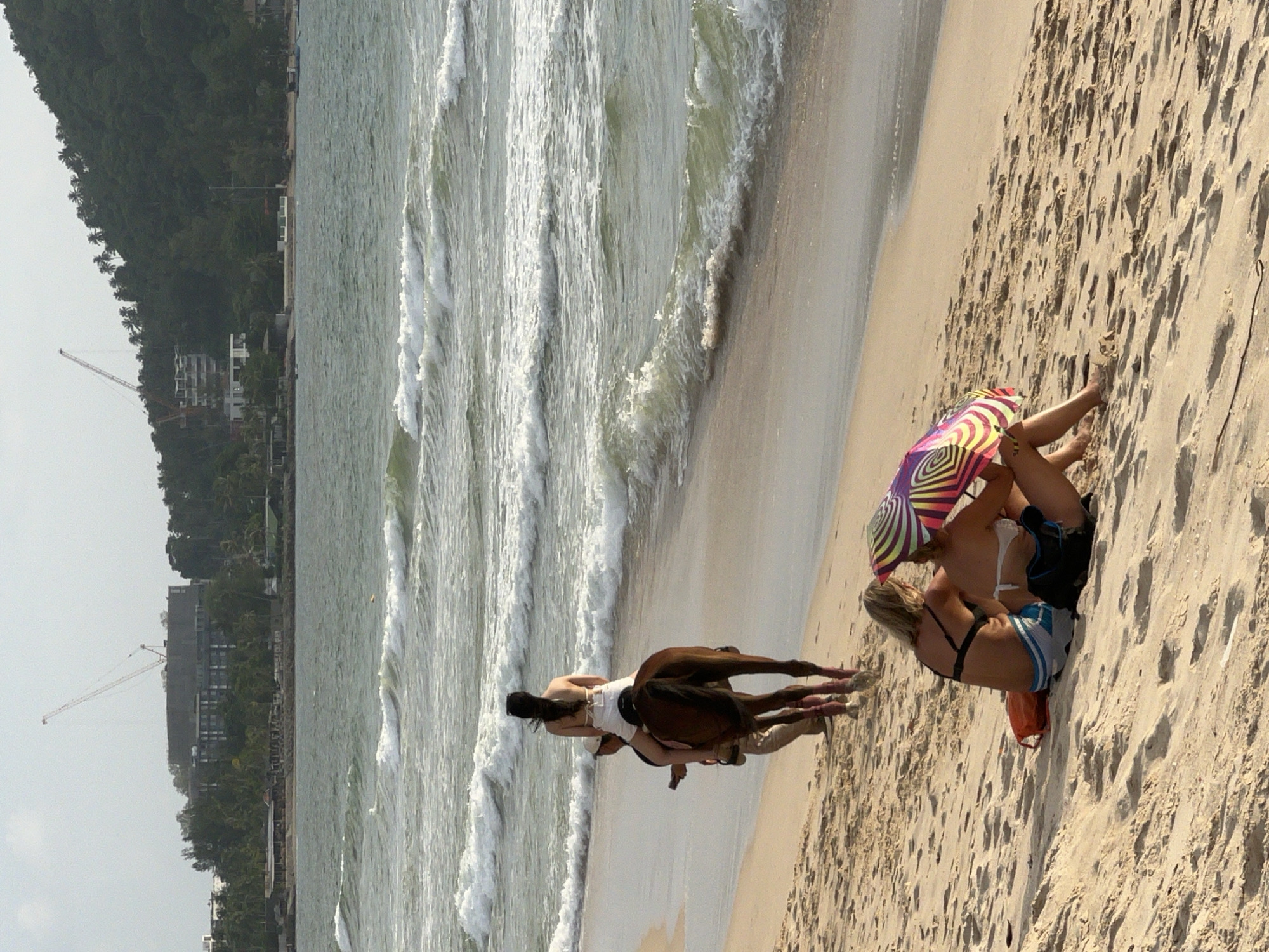 In Choeng Thale, Thailand, on a sandy beach, a person rides a brown horse along the shoreline. The rider, wearing a white top and shorts, faces away from the camera. Accompanying the horse on the sand is a person, likely a guide, walking beside it and holding a phone. In the foreground, two individuals sit on the beach, facing each other. One is wearing a black bikini top and striped shorts, while the other is in a white bikini top and bottoms, holding a colorful, striped umbrella. An orange bag and a black backpack are visible near them on the sand. The ocean with waves stretches to the horizon, and in the background, buildings and a construction crane are visible on a tree-covered hillside.