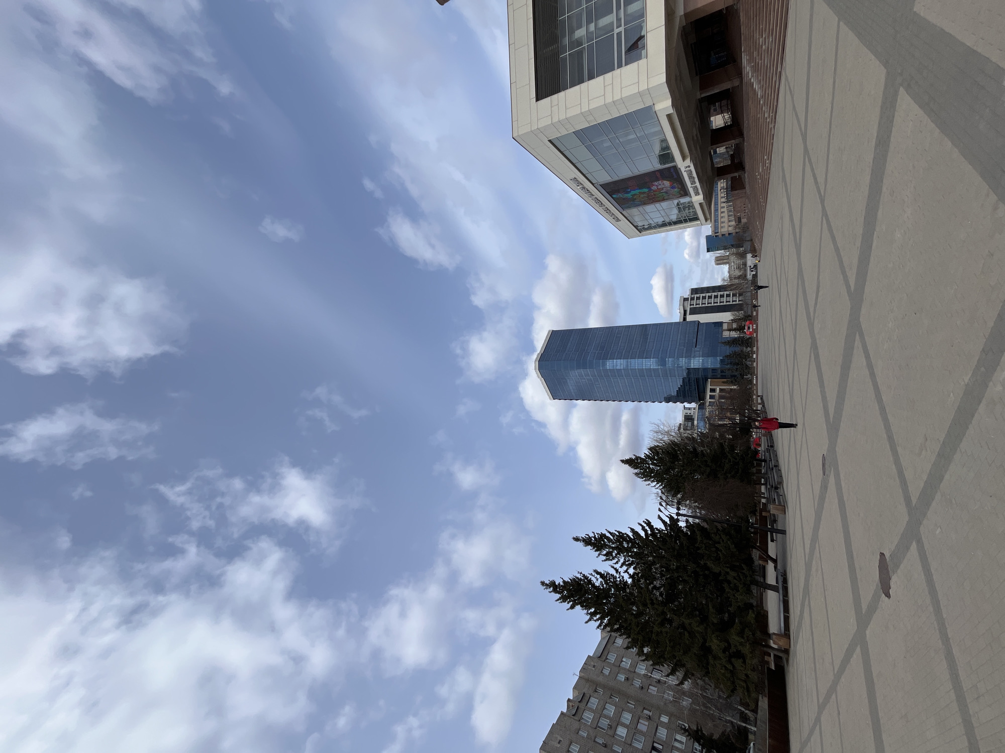 A low-angle, horizontally-oriented photograph shows a cityscape on a cloudy day. A large, modern glass skyscraper stands tall in the center of the frame, reflecting the sky. Adjacent to the skyscraper is a multi-story building with a geometric, tiled façade. A lower, Soviet-era apartment building sits at the base of the skyscraper. The foreground is dominated by a concrete plaza with a grid pattern. Trees line the plaza. A person in a red coat walks along the plaza. The sky is blue with many scattered clouds. The location is likely in the city center of Krasnoyarsk, Russia, indicated by the architectural styles and urban layout.
