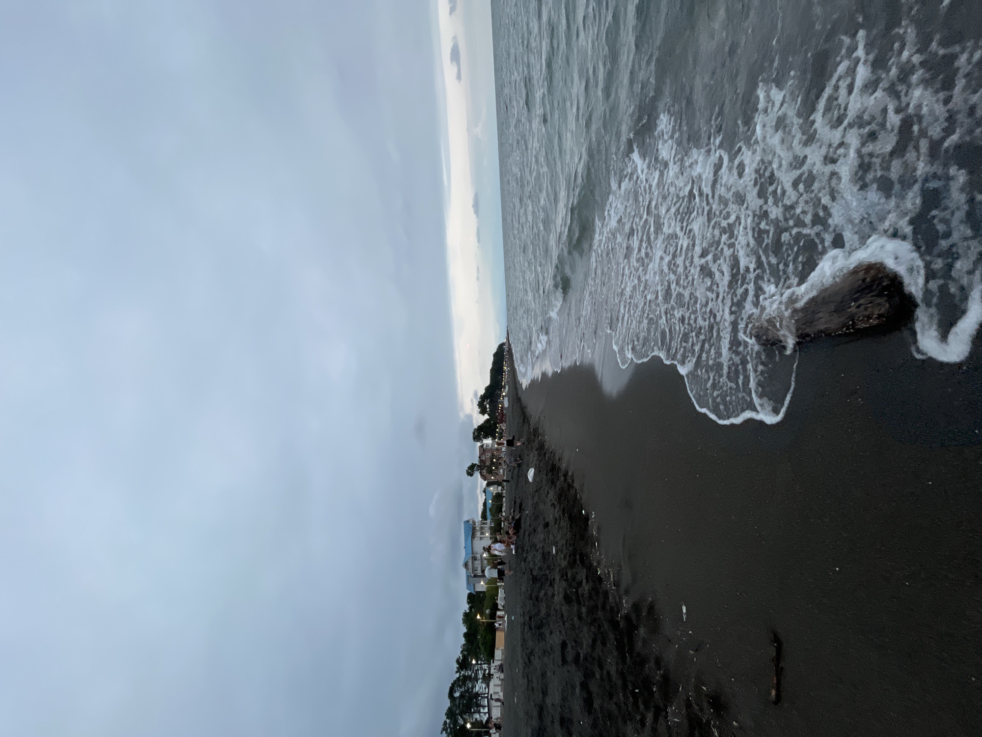 The image depicts a dark sand beach in Ureki, Georgia, under an overcast sky. Gentle waves with white foam are lapping the shore. A piece of driftwood lies partially submerged in the water near the beach. In the background, several buildings, some with blue roofs, are visible behind a line of trees and streetlights. A few people are scattered along the beach and boardwalk, some walking and others gathered in small groups. The overall scene suggests a quiet evening at the resort town.
