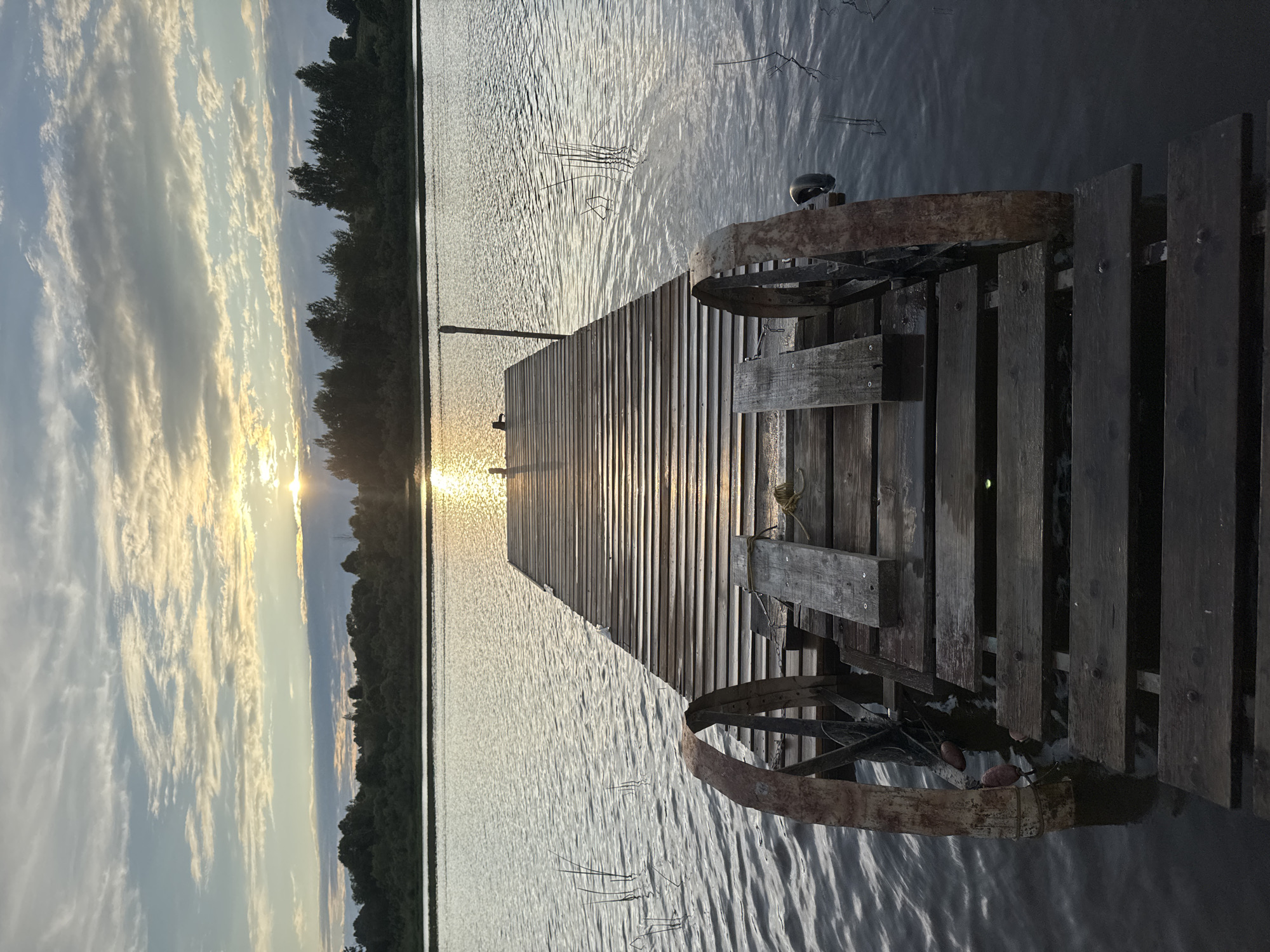 An eye-level, full shot captures a weathered wooden pier extending into a calm body of water, likely a lake or river, near Sitno, Russia. The sun, partially obscured by clouds, casts a warm glow across the scene, creating a shimmering reflection on the water's surface. Tall, dark trees line the opposite bank, silhouetted against the sky. The pier itself appears aged, with worn wooden planks and two large, rusty metal wheels situated on either side of its entrance. The water is slightly rippled, and reeds or tall grass poke out from beneath its surface. The overall atmosphere is serene and tranquil, bathed in the soft light of either sunrise or sunset. There are no people or animals visible in the frame.