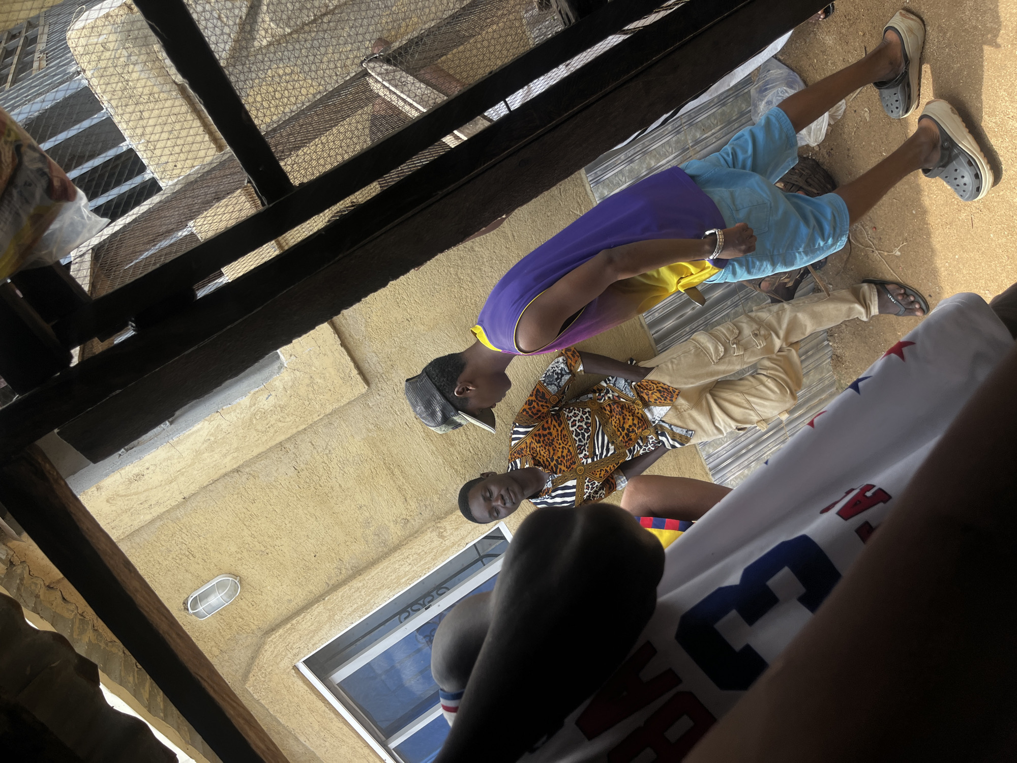 In Rafin Par, Nigeria, two young men stand outdoors under an overhang with a corrugated metal roof and wooden beams. The man on the left, wearing a patterned shirt with leopard print and black and white stripes, and khaki cargo pants, leans against a textured yellow wall. He is looking down, his expression unreadable. To his right, another young man in a purple and yellow tank top and light blue shorts stands facing away from the camera. He wears a grey baseball cap and a silver bracelet on his left wrist. The ground beneath them is dirt. In the foreground, a partially visible person wearing a white t-shirt with large red and blue lettering, including the number "3", is in focus. The background shows a metal fence with diamond-shaped openings and more of the yellow building.