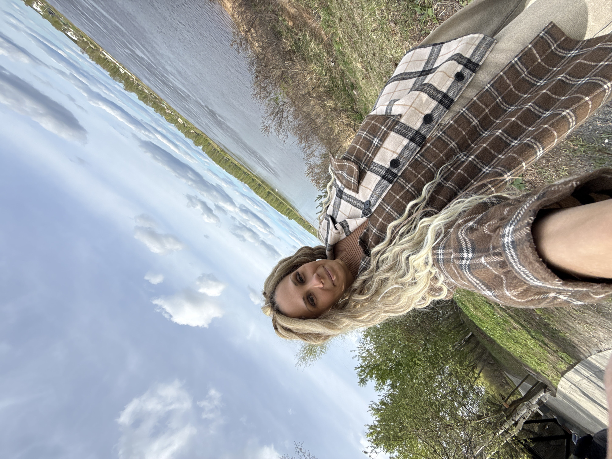 A medium, eye-level shot shows a woman with long, blonde, wavy hair wearing a brown and beige plaid jacket, seated outdoors in Заволжье, Russia. She gazes towards the camera with a slight smile. Behind her, a body of water stretches to the horizon. The sky above is lightly clouded. To her right, a line of trees and greenery extends along the edge of a road.