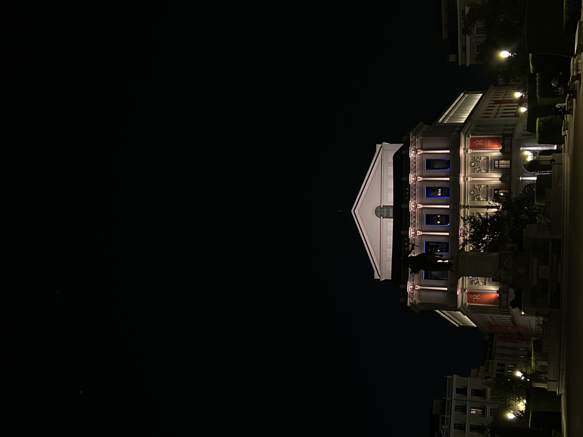 The image shows the Teatro Real in Madrid, Spain, at night. The building is illuminated with white and blue lights. The facade of the theater is visible, with its columns, windows, and a sign with the letter "R." In front of the theater, there is a statue. In the background, other buildings and streetlights can be seen. The sky is dark.
