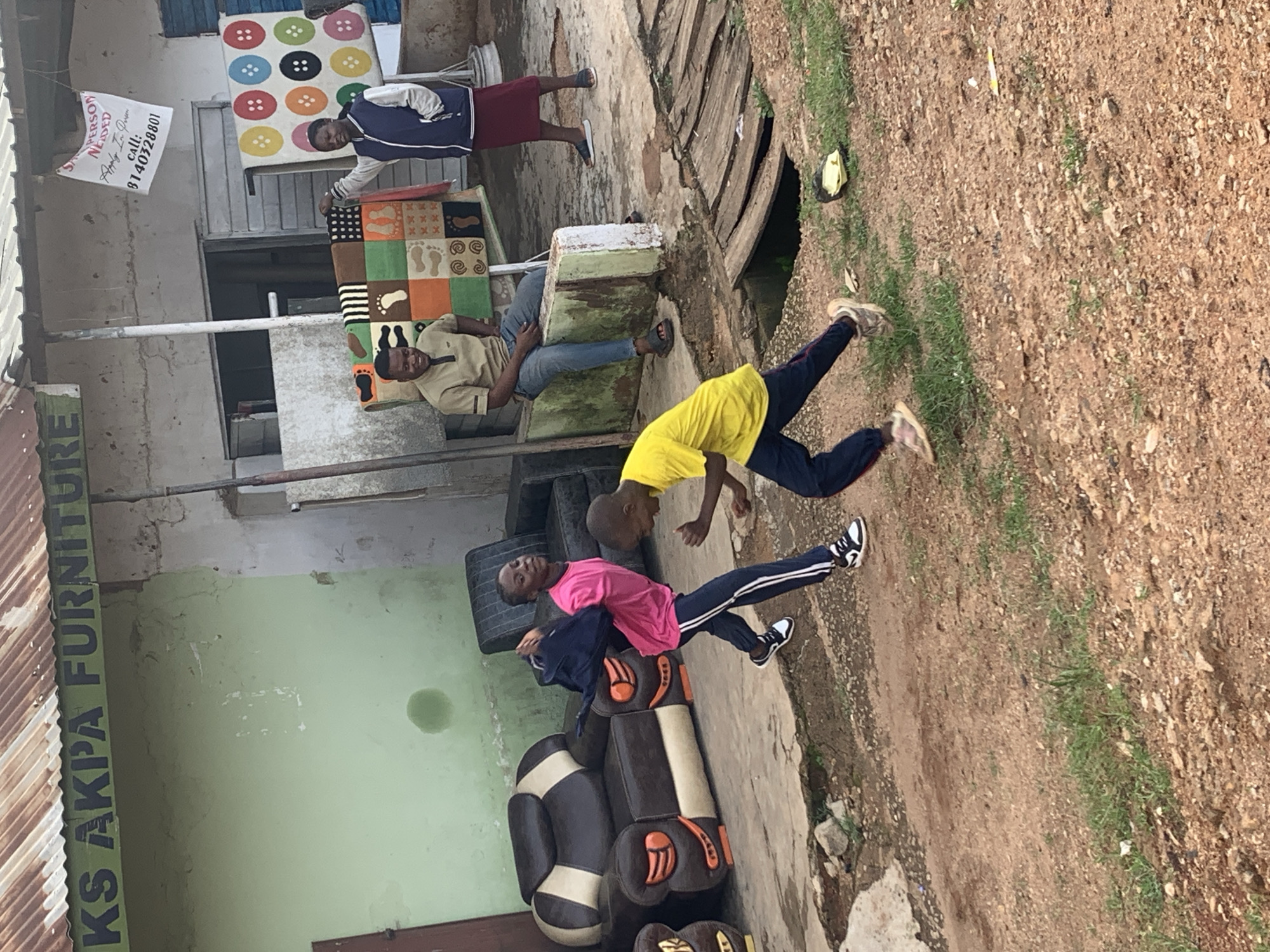 The image depicts a scene outside what appears to be a furniture shop in Jos, Nigeria. Several people are present, including three young boys and two adults. One adult, a man, is seated and looking towards the camera. Another adult, a woman, is standing and looking away from the camera.
The central activity involves the three boys, one of whom is dressed in a pink t-shirt and dark pants, is lying on a sofa. Another boy in a yellow t-shirt and dark pants is in a playful, energetic pose, appearing to be either running or jumping. The third boy, partially visible, is also interacting in the foreground. The setting includes a variety of objects, such as sofas, tires stacked up, and colorful rugs displayed on a wall. One rug features a pattern of colorful circles, while another has a mosaic of footprints and other graphic designs. A white sign hangs on the wall with text that reads "SALES PERSON NEEDED Apply 'LE' Seven call: 8140328801." The furniture shop appears to be named "AKS AKPA FURNITURE" as indicated by text on a green banner.
The environment suggests an outdoor, casual setting, possibly a commercial area with limited infrastructure. The ground is a mix of dirt and some patchy grass, with a large opening in the ground visible in the background. The lighting suggests it is daytime, likely under natural sunlight, though the exact time of day and weather conditions are not explicitly discernible. The overall mood seems relaxed and informal, with the children engaged in play.