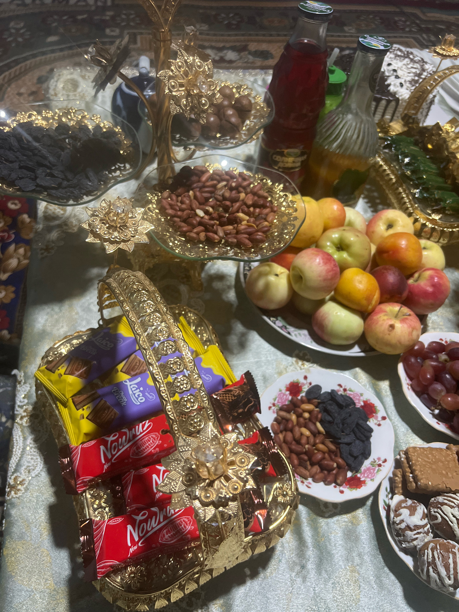 An overhead, slightly angled medium shot captures a table laden with a variety of food items, likely set for a celebration. The table is covered with a light blue and white patterned cloth. Dominating the center is a golden, ornate, multi-tiered stand holding various dried fruits, nuts, and possibly chocolates on different levels. To the left, a glass dish contains dark, dried grapes. Below this, a golden woven basket is filled with individually wrapped chocolates, some in yellow and purple packaging with "Laico" branding, and others in red packaging with "Nowruz" branding. To the right of the basket, a white plate displays a mix of peanuts and dark, dried grapes. Further to the right, a pile of assorted apples of various colors is arranged on a floral-patterned plate. Below the apples, a white dish holds a cluster of dark red grapes. In the bottom right corner, another white plate contains chocolate-covered biscuits and spherical chocolates drizzled with white icing. Two glass bottles, one containing a red liquid and the other a golden liquid, stand on the table behind the golden stand. A green straw is visible between the bottles. The background is blurred but appears to be a patterned rug. There are no people or animals visible in the image. The overall impression is one of abundance and festive preparation.