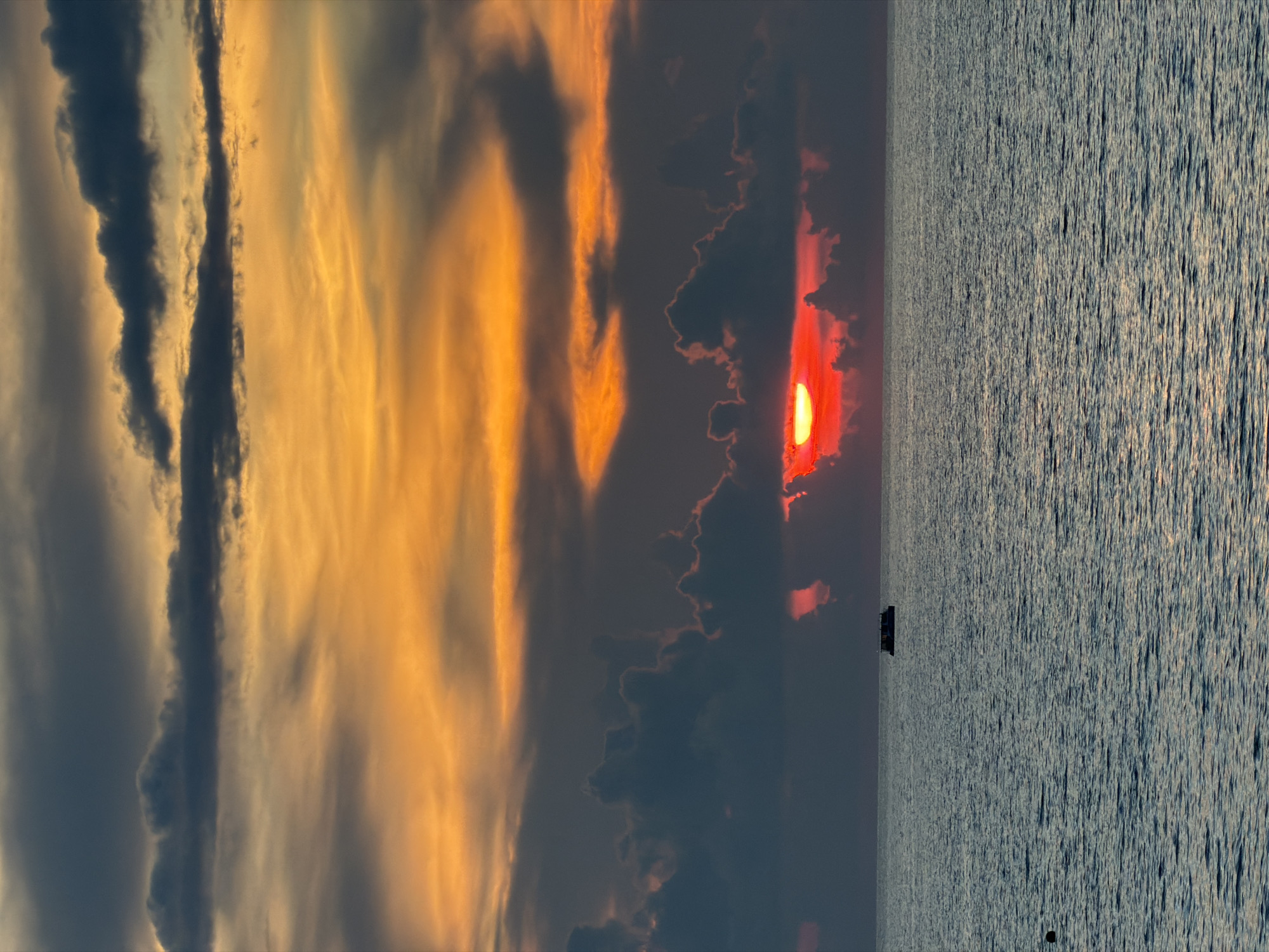 A sunset over the ocean is captured in this vertical, medium-shot photograph. The sun, a brilliant red orb, is partially obscured by dark, heavy clouds near the horizon, casting a warm orange and yellow glow through the sky. The ocean in the foreground is a deep blue, with subtle ripples reflecting the twilight. A small, dark boat with a light-colored superstructure is visible on the water, centered horizontally and positioned in the lower third of the image. The scene is tranquil, with no visible people or further activity. The location is listed as Unknown, Unknown.