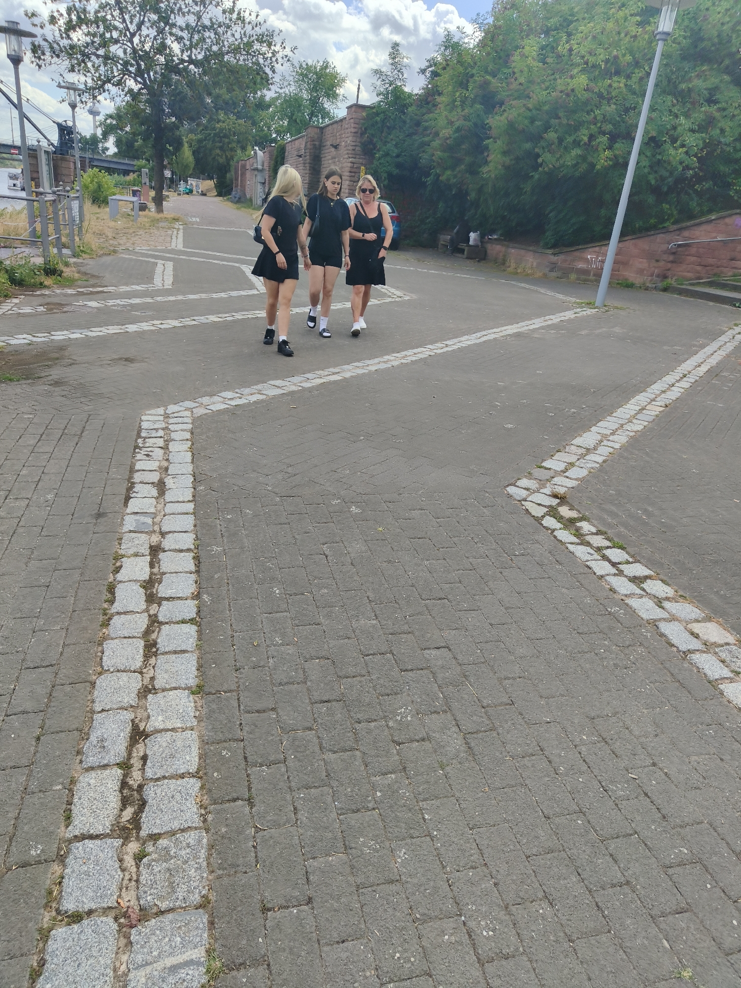 Three women are walking on a paved pedestrian path in Mannheim, Germany. The woman on the left has blonde hair and is wearing a black dress and black shoes. The woman in the middle has brown hair and is wearing a black top and black shorts. The woman on the right has blonde hair and sunglasses, and is wearing a black dress and white sneakers. The path is made of gray bricks with decorative lines of smaller stones. Trees and a stone wall line the right side of the path. In the background, there are more trees, a lamppost, and some buildings. The sky is partly cloudy.