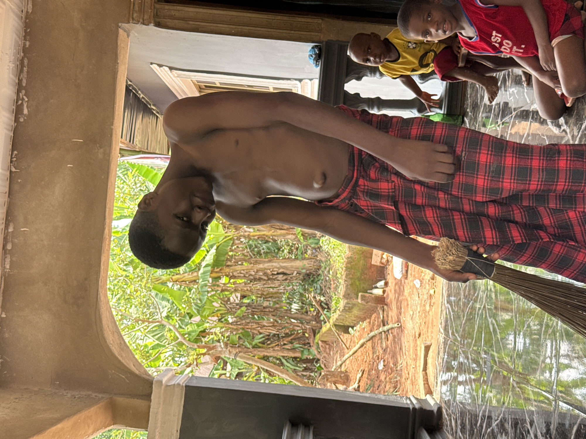 A vertical shot captures a domestic scene in Eziudo, Nigeria, featuring a young male in the foreground, shirtless and wearing a red and black plaid wrap around his waist. He holds a natural fiber broom in his right hand, poised as if sweeping or about to sweep, and looks towards the camera with a neutral expression.
To his right and slightly behind, three young children are visible. The most prominent child wears a red and white t-shirt with the partial text "ST DO" (likely "JUST DO IT") and gazes directly at the camera with a faint smile. Another child, wearing a yellow t-shirt, also looks towards the camera, while a third is partially obscured.
The setting appears to be a covered indoor or semi-outdoor area, indicated by a light-colored, reflective tiled floor and a prominent archway. Beyond the archway, a bright outdoor environment is visible, characterized by vibrant green tropical foliage, possibly banana leaves, and bare tree branches. The ground outside is reddish-brown dirt, with scattered debris including a few empty bottles and fallen branches. The scene is brightly illuminated by natural daylight, suggesting a sunny day. The overall impression is one of everyday life in a rural or semi-rural Nigerian community.