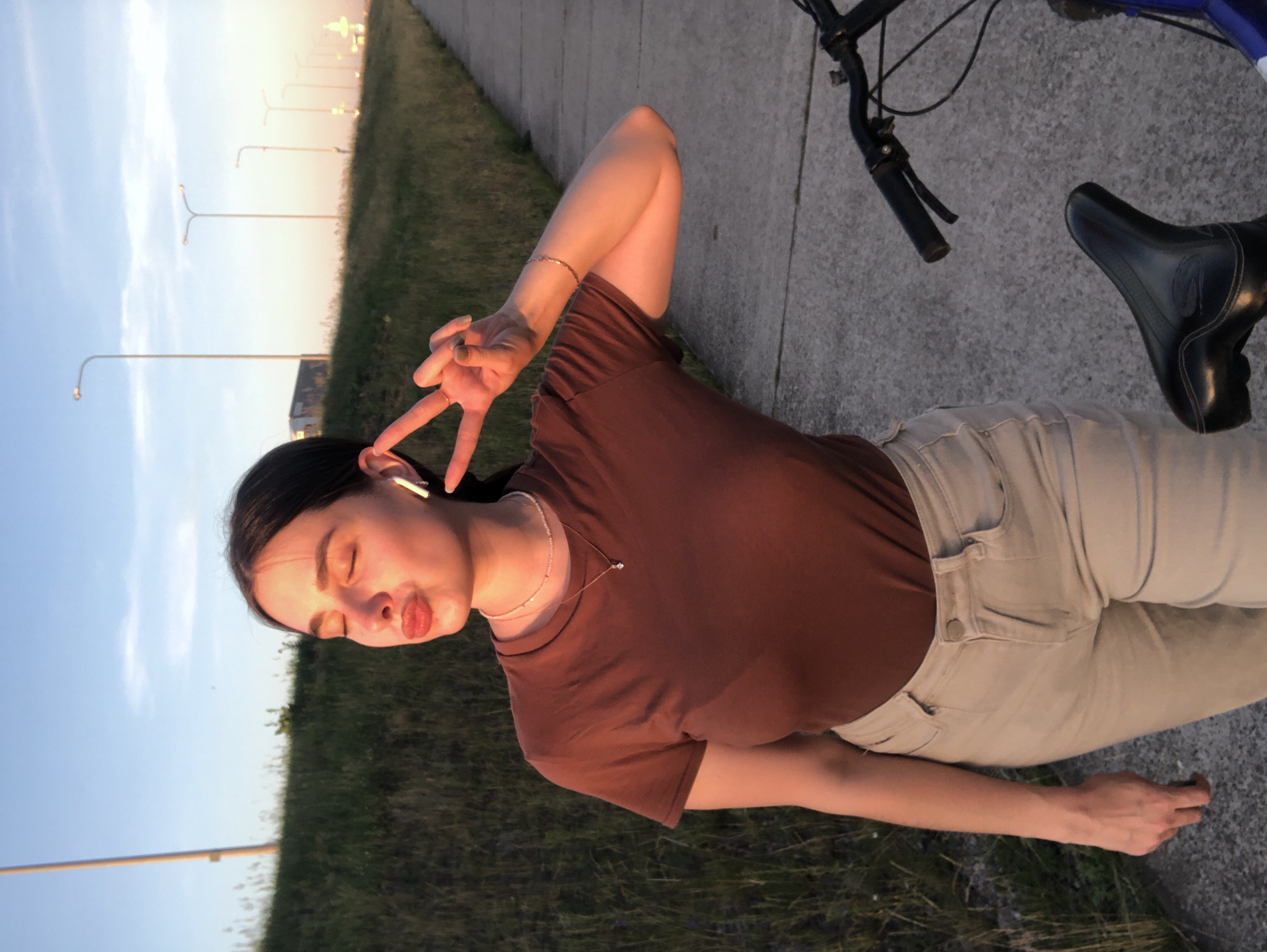 In this vertically oriented, wide shot taken outdoors, a young woman is depicted lying down on a paved surface, possibly a sidewalk or bike path, with a grassy area to her left. The woman has dark hair pulled back and is wearing a brown t-shirt and light-colored pants. Her eyes are closed, and she is puckering her lips, making a "kiss" gesture with her mouth. Her right hand is raised, with two fingers extended in a peace sign, and she is wearing a bracelet on her left wrist and an earbud in her right ear.
Beside her, a bicycle is visible, with its handlebars and seat in the frame. The setting appears to be a suburban or semi-rural area, with a row of streetlights stretching into the distance along the edge of the paved path. The sky is a pale blue, suggesting it is either early morning or late afternoon, with the warm light casting a soft glow on the scene, indicating it is either sunrise or sunset. The overall mood is relaxed and playful. There is no visible text in the image.
