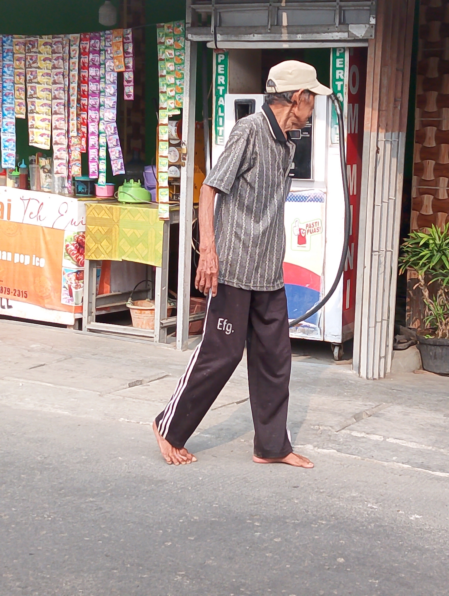 An elderly Indonesian man wearing a grey striped shirt, black track pants with white stripes, and a light-colored baseball cap is walking barefoot on a paved street in Karawang, Indonesia. The man is walking from left to right, with his head turned to his right. He is in front of a small shop that appears to be selling snacks and possibly gasoline. The shop has a green awning and a variety of colorful products displayed on shelves. Behind the man, a white gasoline pump with red and blue accents stands near the shop entrance. A potted plant is visible to the right of the gasoline pump. The scene suggests a typical street view in a commercial area of Karawang.