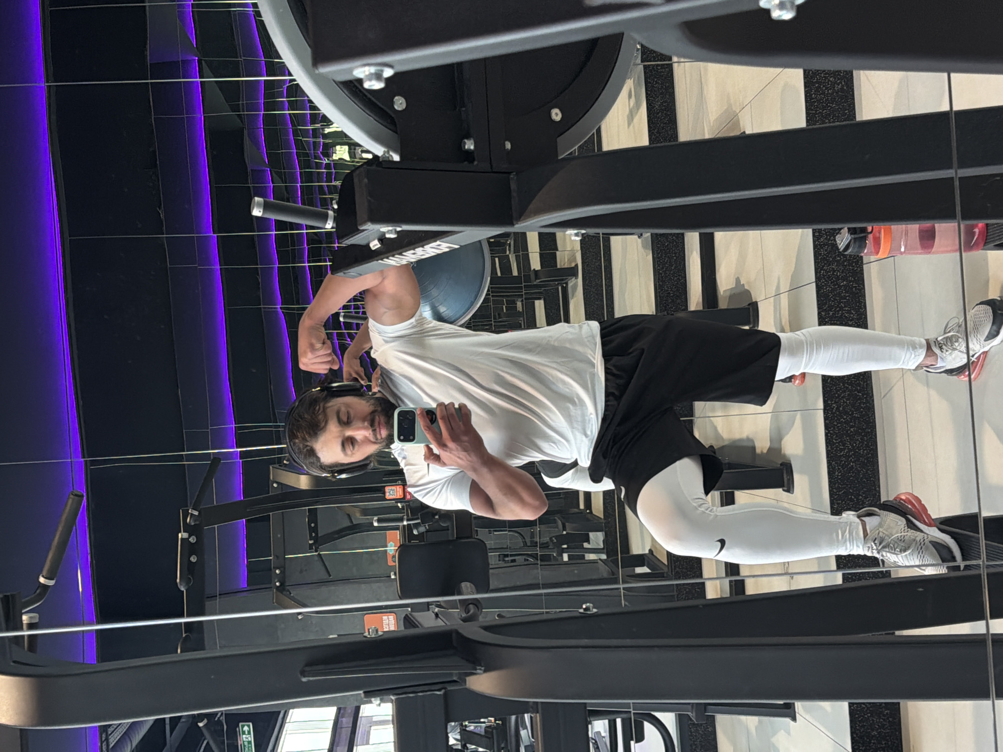 A man is taking a selfie in a gym mirror in Krasnoyarsk, Russia. He is wearing black shorts, a white t-shirt, white leggings, and sneakers. He has headphones on and is flexing his bicep. The gym has black equipment, purple lighting, and a black and white striped floor. A water bottle is near the edge of the shot.