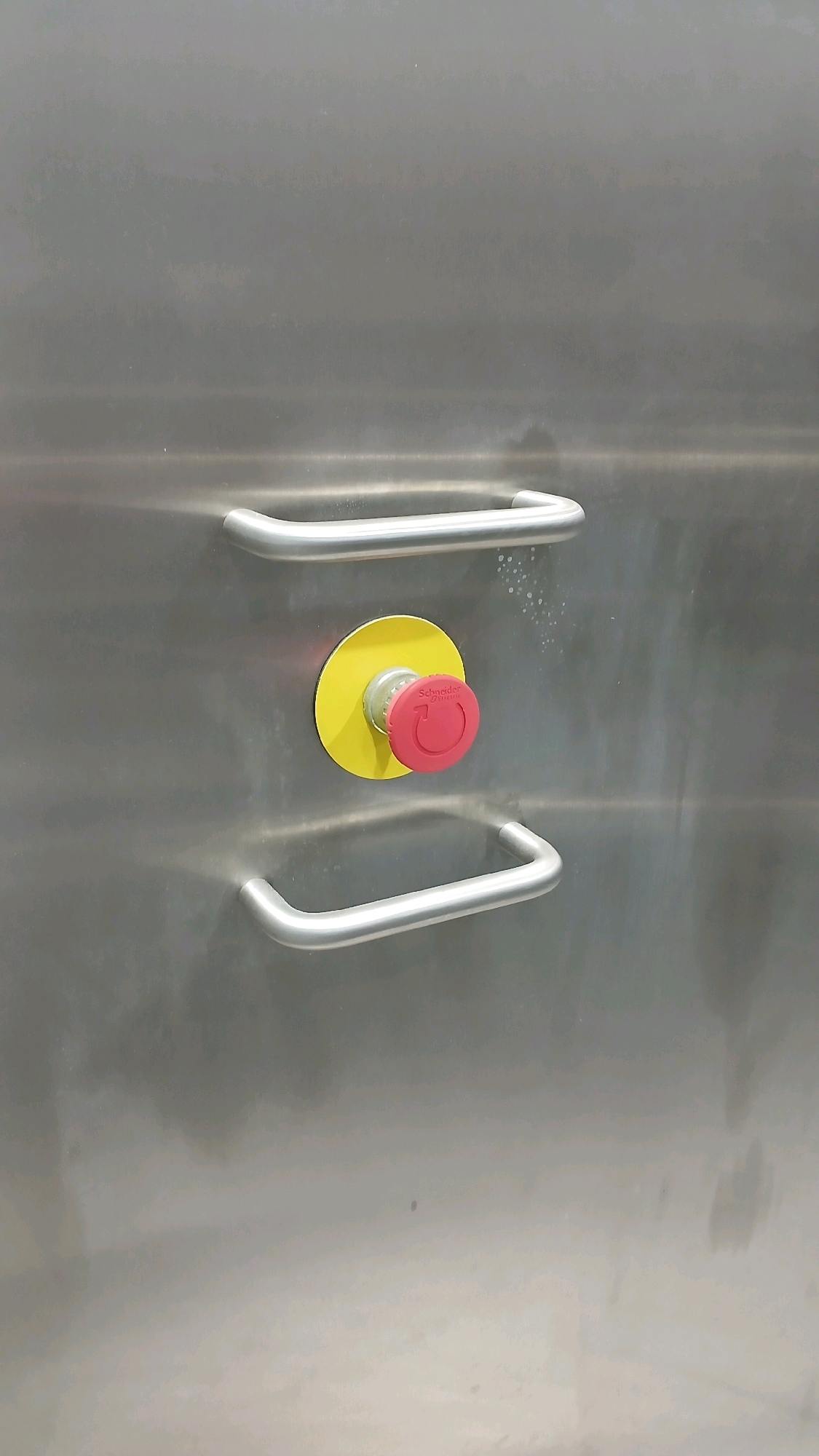 The image shows a close-up, eye-level, vertical shot of a stainless steel surface with two brushed metal handles and a red emergency stop button with a yellow background. The button is circular with a mushroom-shaped head and has the Schneider Electric logo visible on it. The surface appears to be the door or panel of a piece of equipment. The lighting is neutral, and there are a few water droplets or smudges on the stainless steel above the button. There are no people or other discernible objects in the immediate vicinity, and the background is a blurry, diffused gray. The scene is forensic and industrial in nature. The location of Gadobangkong, Indonesia is not visually represented in the image.