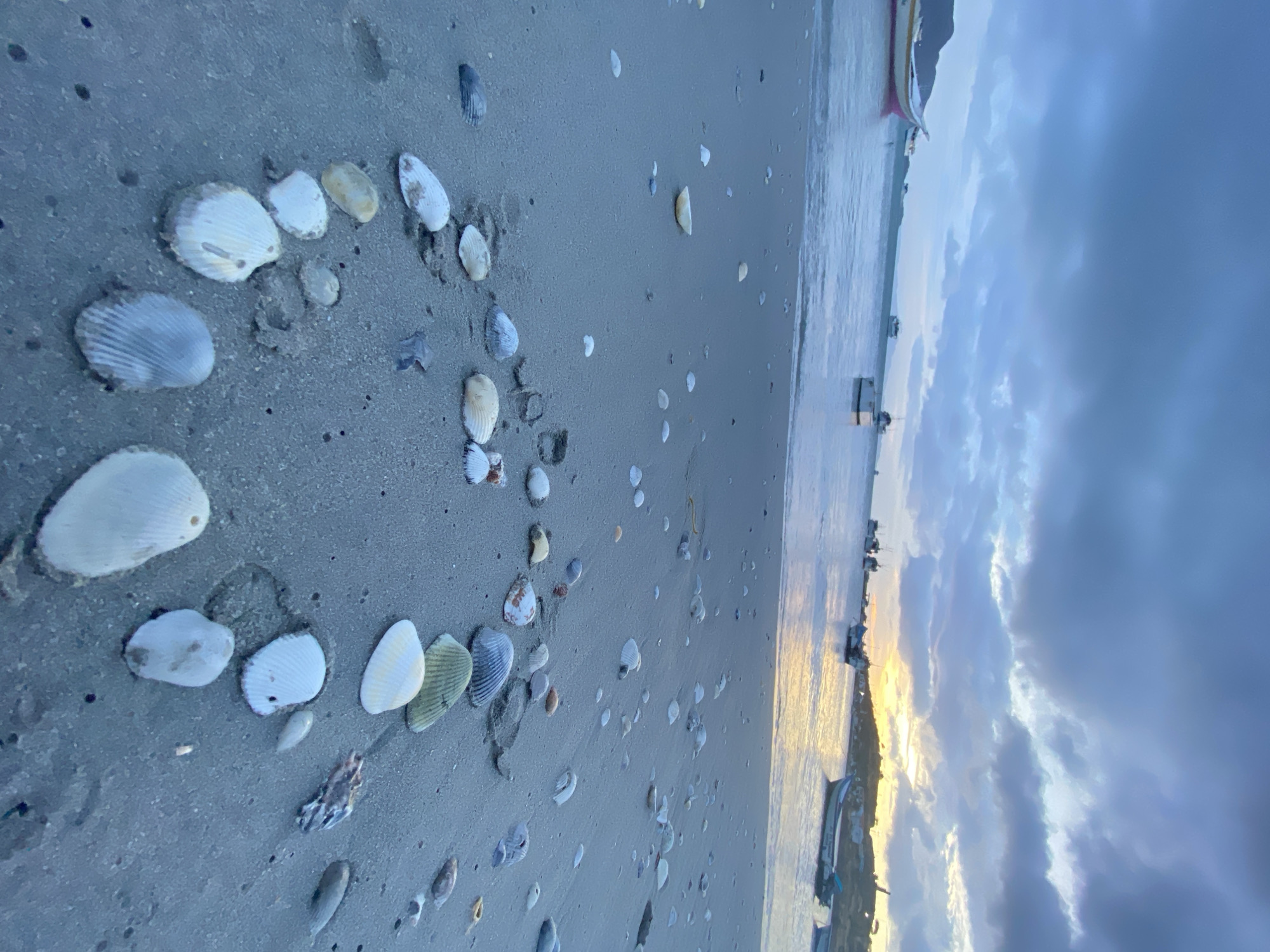 The image captures a scene at Playa Caribe in Juan Griego, Venezuela. A partial circle of seashells is arranged on the sandy beach. The shoreline is visible in the background, with boats anchored in the water. The sky is overcast with dark clouds, with a glimpse of sunset light on the horizon. No people are visible in the shot.