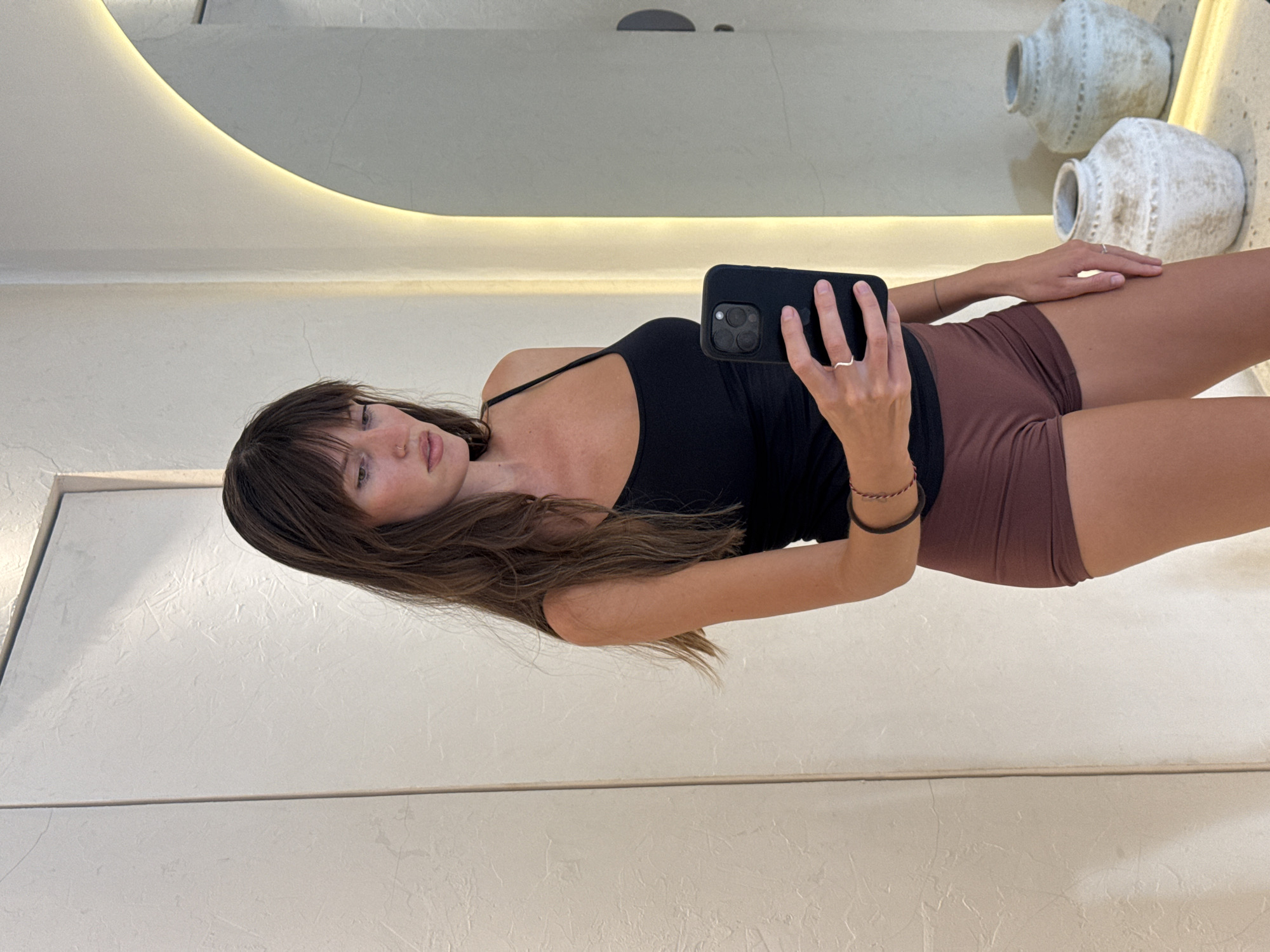 A woman in a black tank top and brown shorts stands indoors, holding a black smartphone in her right hand to take a selfie in front of a mirror. Her left hand rests on her right thigh. She has long, brown hair with bangs. In the background, there are decorative pots on a shelf. The image was taken in Cemagi, Indonesia.