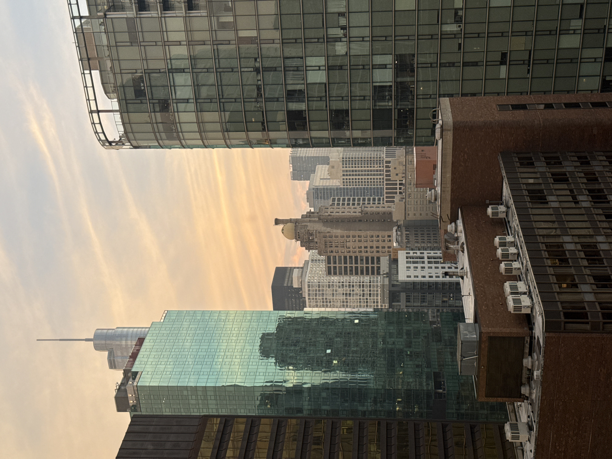 This image depicts a view of the Chicago skyline during a sunset. The sky is filled with soft, wispy clouds painted in shades of orange and yellow, suggesting the golden hour. In the foreground, several buildings are visible. On the left, a dark-colored building with horizontal windows is partially shown. Adjacent to it is a tall, striking building with a facade of greenish-blue glass, reflecting the surrounding structures and the sky. Further to the right, a curved, modern building with a glass exterior dominates the mid-ground, its many windows revealing interior lights and architectural details. Behind these, a cluster of other skyscrapers rises, including a tall building topped with a spire, and a building with a distinctive golden dome. The lower portion of the image shows the rooftops of closer buildings, characterized by brickwork and various mechanical units, likely HVAC systems, indicating an urban setting. The overall impression is one of a dense cityscape bathed in the warm, diffused light of dusk.