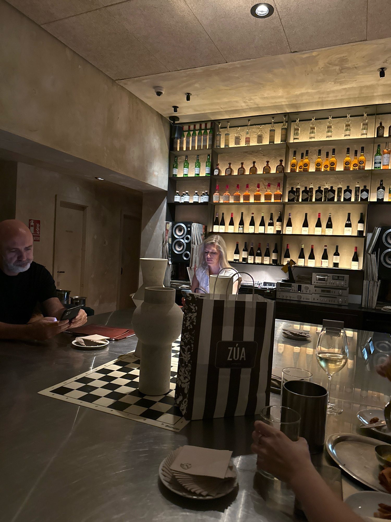 The image depicts an interior scene at what appears to be a bar or restaurant in Madrid, Spain. In the foreground, a man with a bald head and a white beard is seated at a metal bar counter, holding a smartphone in his left hand. In front of him, a small white plate with napkins and a red folder are visible. To his right, on the counter, are two decorative vases. A chessboard is laid out on the counter behind the vases.
The bar area features extensive shelving filled with various bottles of alcohol, illuminated from behind. A woman with blonde hair is seated behind the bar, looking at a screen. To her left, a shopping bag with the text "ZÚA" is placed on the counter. A glass of white wine sits in front of a person whose hand is reaching for a drink.
The environment is dimly lit, with spotlights on the ceiling and accent lighting behind the shelves of bottles. The walls are a muted beige color, and the ceiling appears to be a textured material. The overall atmosphere suggests a sophisticated and contemporary establishment.