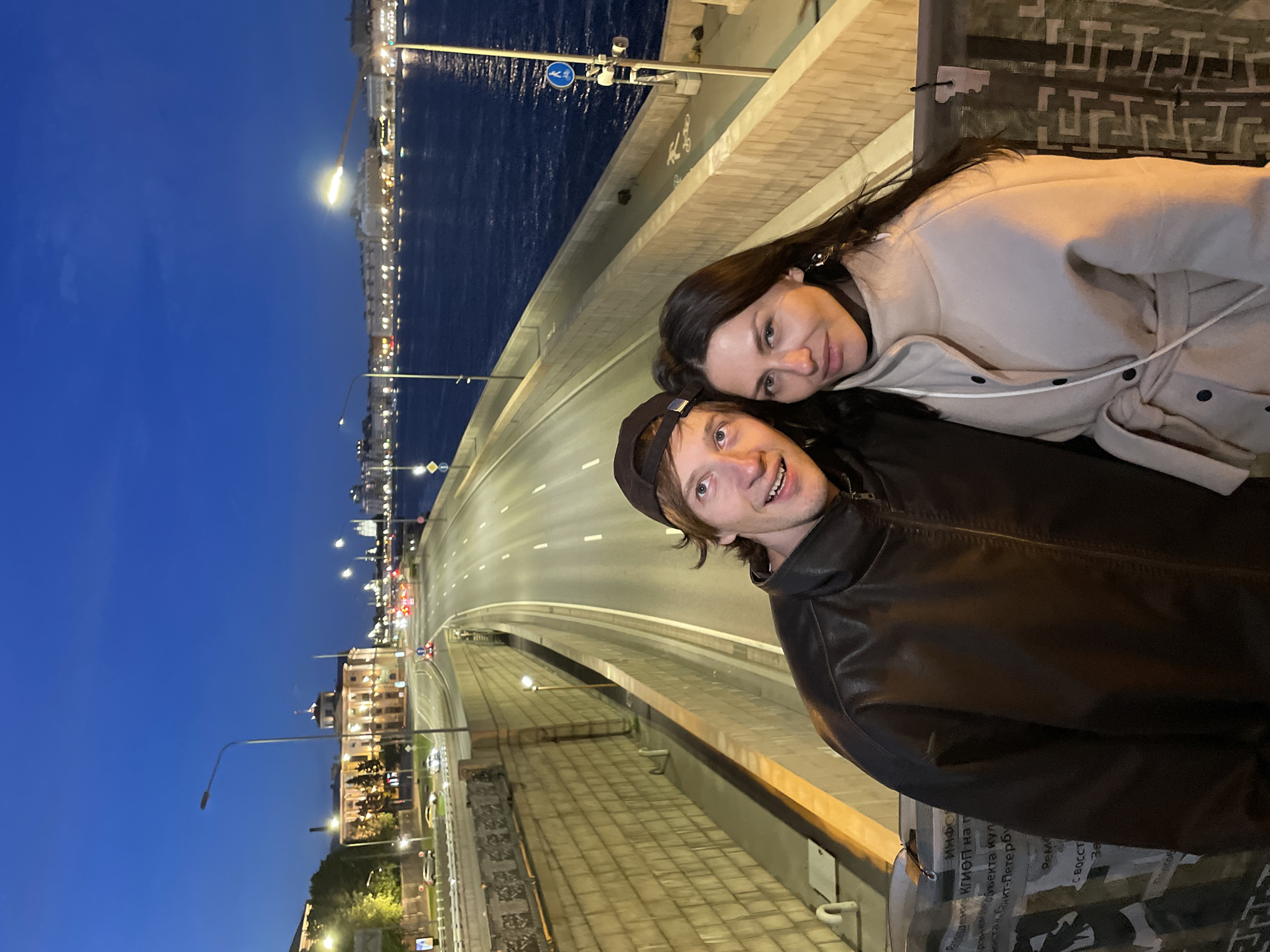 A couple poses for a selfie on a bridge at dusk in Saint Petersburg, Russia. The man, wearing a brown leather jacket and a baseball cap, smiles broadly, while the woman, in a cream-colored coat, looks directly at the camera. In the background, a wide road with traffic lights and vehicles stretches towards a cityscape illuminated by lights. A dark river, likely the Neva River, runs alongside the road, reflecting the urban lights. The sky is a deep twilight blue. The bridge has stone walls and streetlights, and there are signs indicating a bike path.