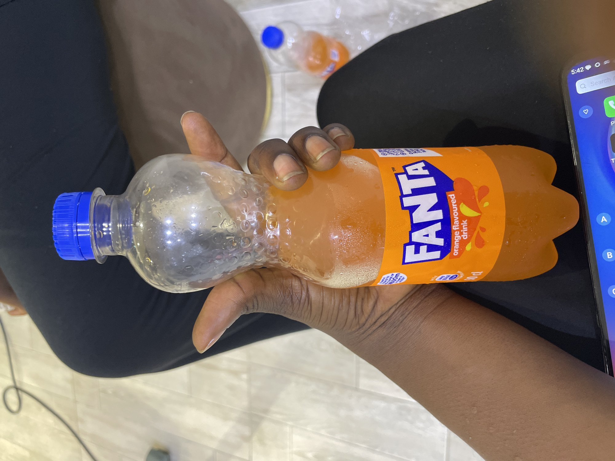 An overhead close-up shot shows a person's hand holding a plastic bottle of Fanta. The bottle is partially filled with orange liquid and condensation. The label on the bottle is visible, displaying the Fanta logo and text that reads "orange flavoured drink." The person's skin tone is dark, and their fingernails appear unpolished. In the background, a person wearing dark trousers is visible, along with a floor that appears to be tiled. Another partially visible bottle of Fanta is on the floor in the background. The overall scene suggests a casual indoor setting.