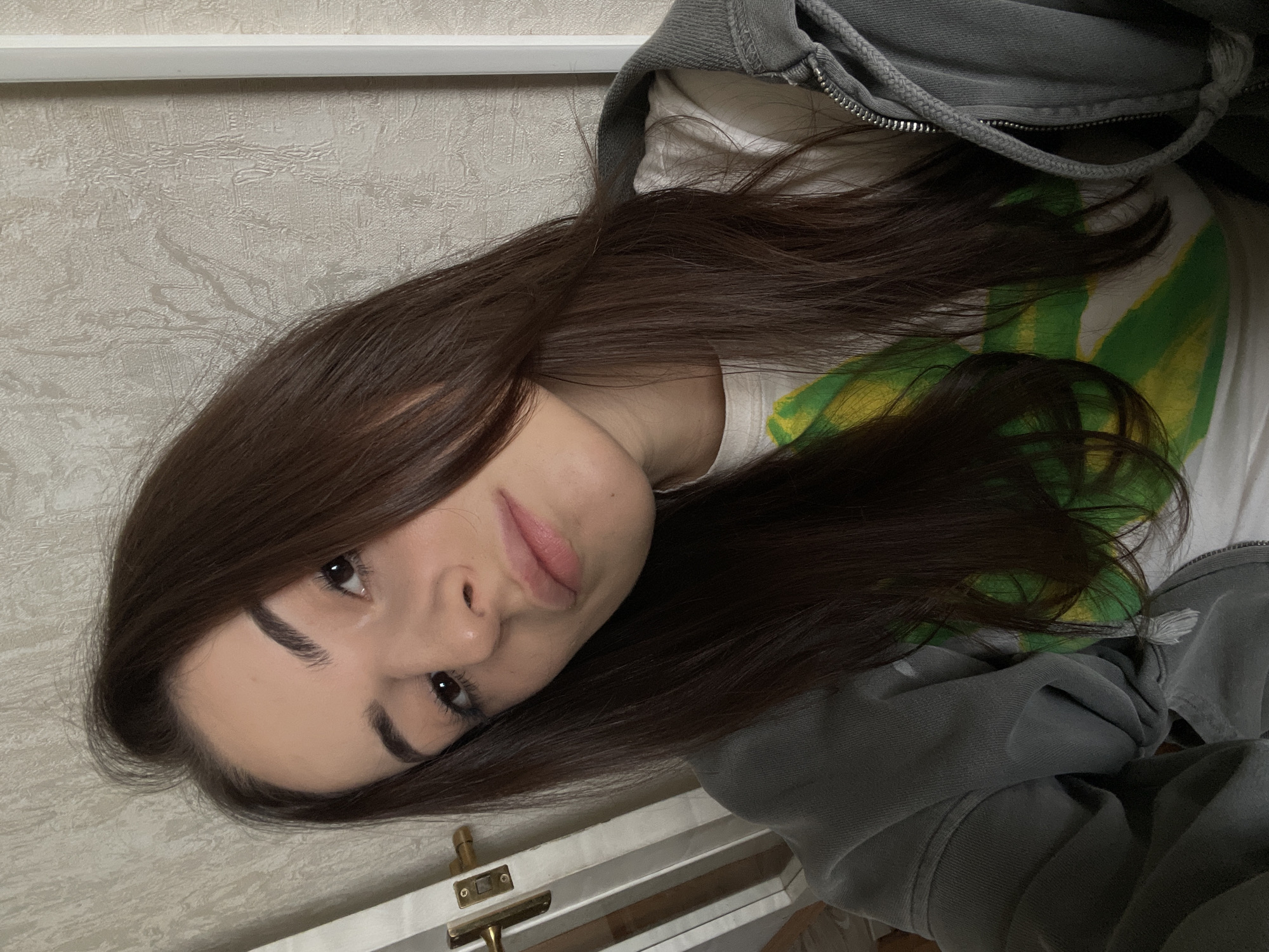 A young woman with long, dark brown hair is visible from the shoulders up, tilted at an angle facing the camera. She has dark eyebrows and eyes, and light-colored skin. She is wearing a white t-shirt with green and yellow graphics, and a grey zip-up hoodie. The background consists of a beige wall with a textured pattern. A white trim is visible at the top of the wall. A door with a gold handle and hinges is visible in the lower left corner of the image. The location is unknown, but based on the prompt is not Russia.