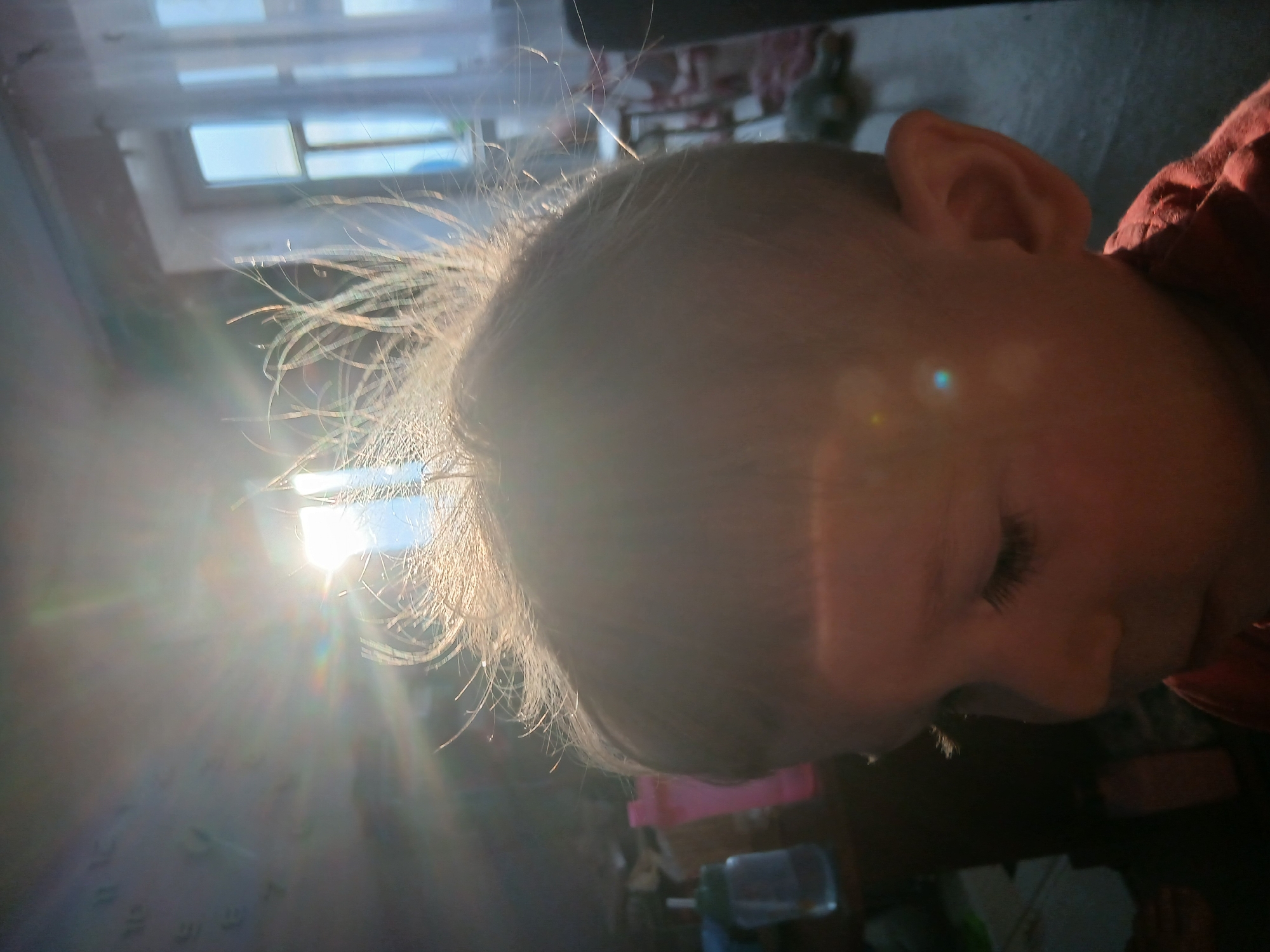 The image depicts a child's face in close-up, seemingly asleep or resting. The child has fair hair, with individual strands highlighted by sunlight. The ear and a portion of the face are visible, along with closed eyelashes. The background is blurred, but a window with a bright light source is evident. Other objects are obscured, but shapes suggest furniture and other room contents. The location is Chaikovsky, Russia, as context is provided. The light is bright, possibly sunlight entering through the window.