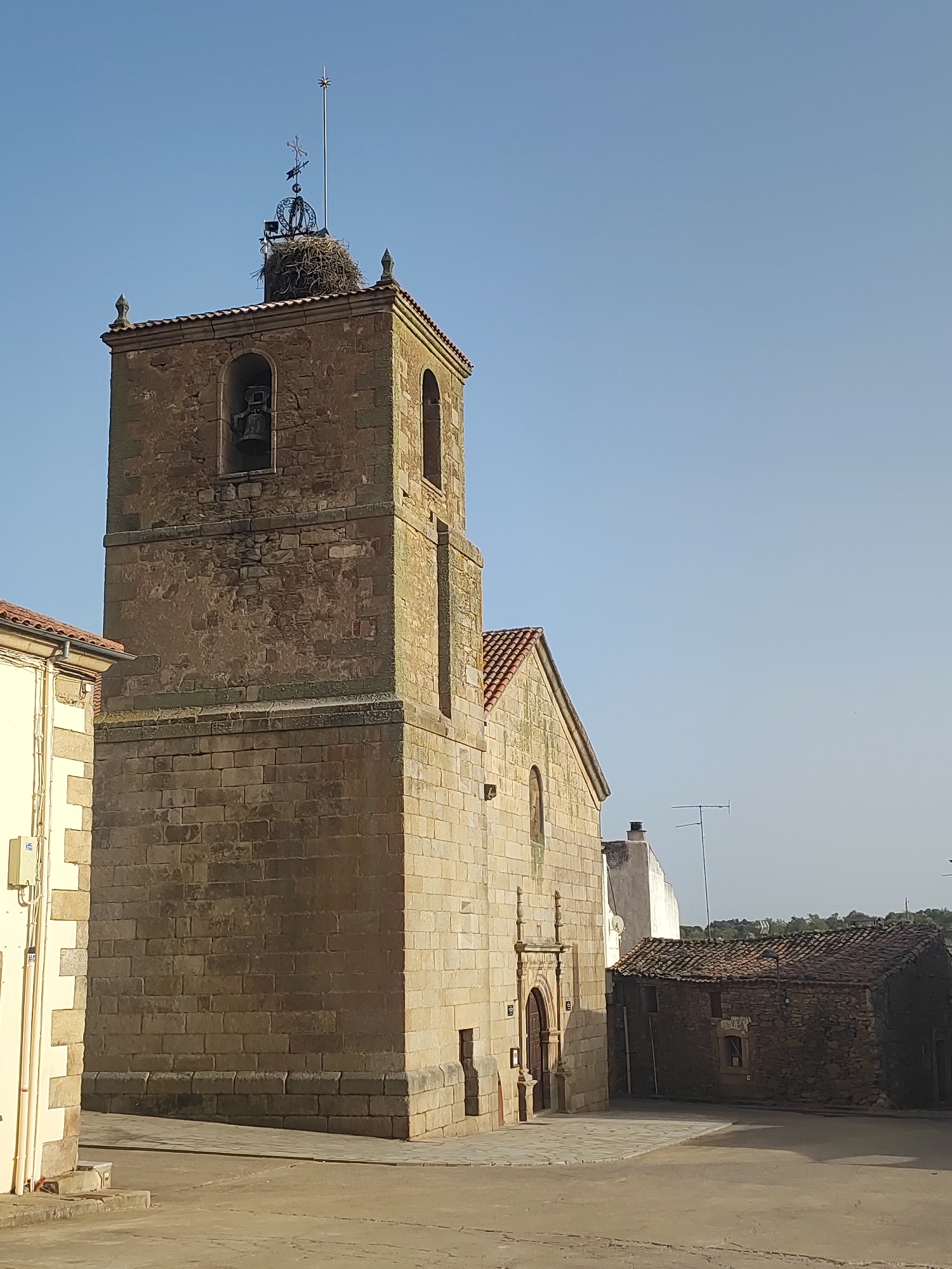 The image captures a stone bell tower with a stork's nest at its summit, located in the city of Sobradillo, Spain. The tower, constructed from large, weathered stone blocks, features arched openings for bells and a decorative element at its apex. Adjacent to the tower is a smaller stone building with a tiled roof, and a portion of a cream-colored building with visible electrical conduits is visible on the left. The ground in the foreground is a paved area, appearing to be a street or plaza. The sky is clear and blue, with the sun casting long shadows. There are no people visible in the scene.