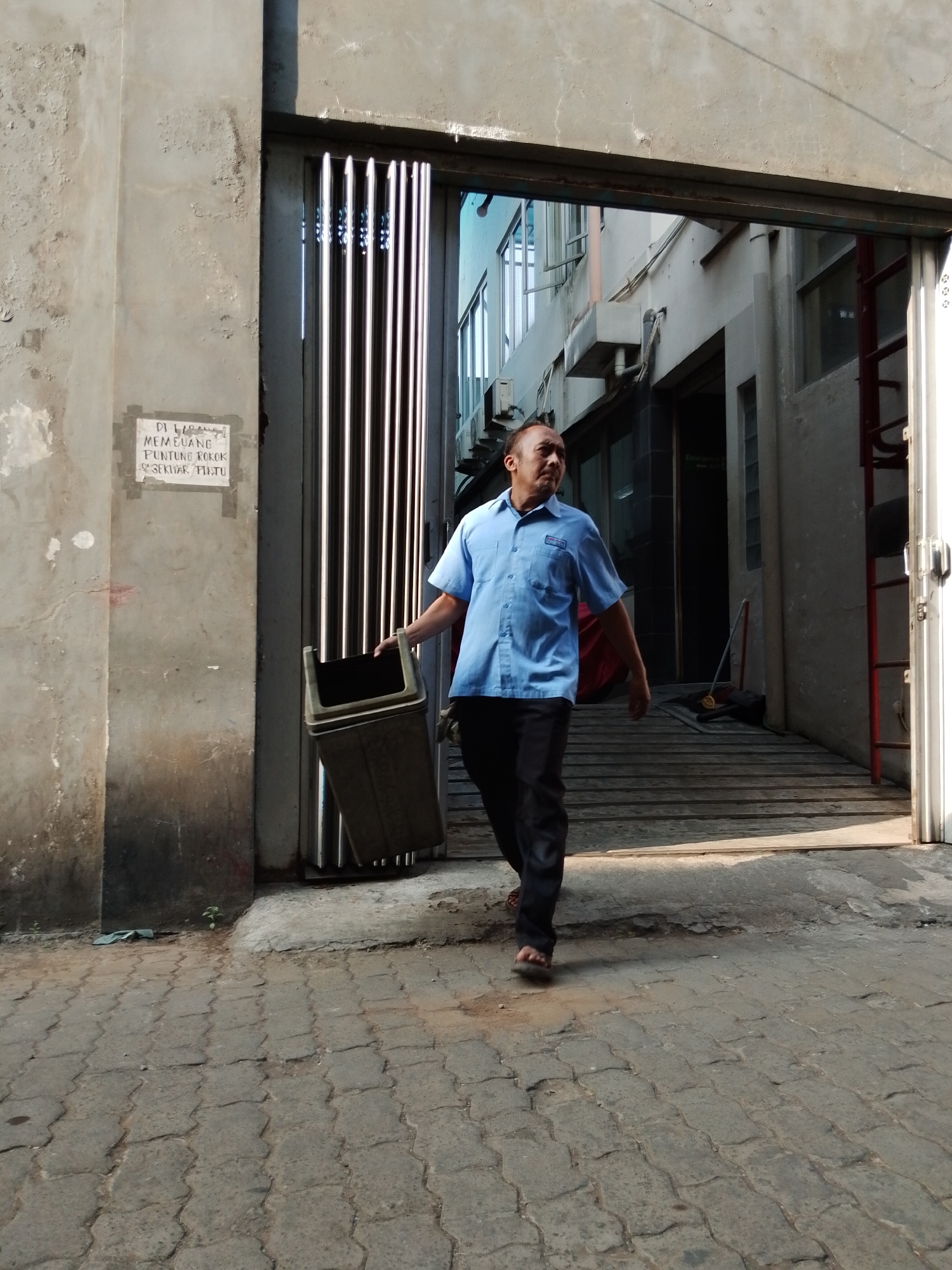 In Pasuruan, Indonesia, a man in a light blue collared shirt and dark pants walks towards the camera, carrying a grey trash bin. He appears to be exiting a building through a wide opening with vertical metal slats on the left side of the frame. The ground is paved with interlocking grey bricks. A sign is visible on the wall to the left of the opening, with text that appears to be in Indonesian. The building interior shows a ramp leading up to a door, and a red ladder is visible on the right side of the opening. The scene is lit by bright sunlight, casting shadows on the ground.