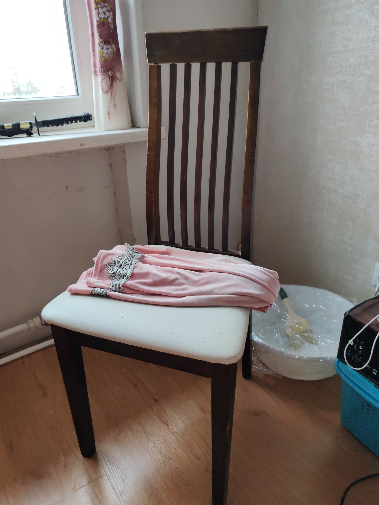A wooden chair with a white cushion has a pink, lace-trimmed garment draped over it. A white bowl covered in plastic wrap and containing a paint brush sits next to the chair, along with a blue plastic basket and a black electronic device. A katana sword rests on the window sill. The location is likely indoors and is probably not possible to locate in Petropavlovsk-Kamchatsky, Russia with the available information.