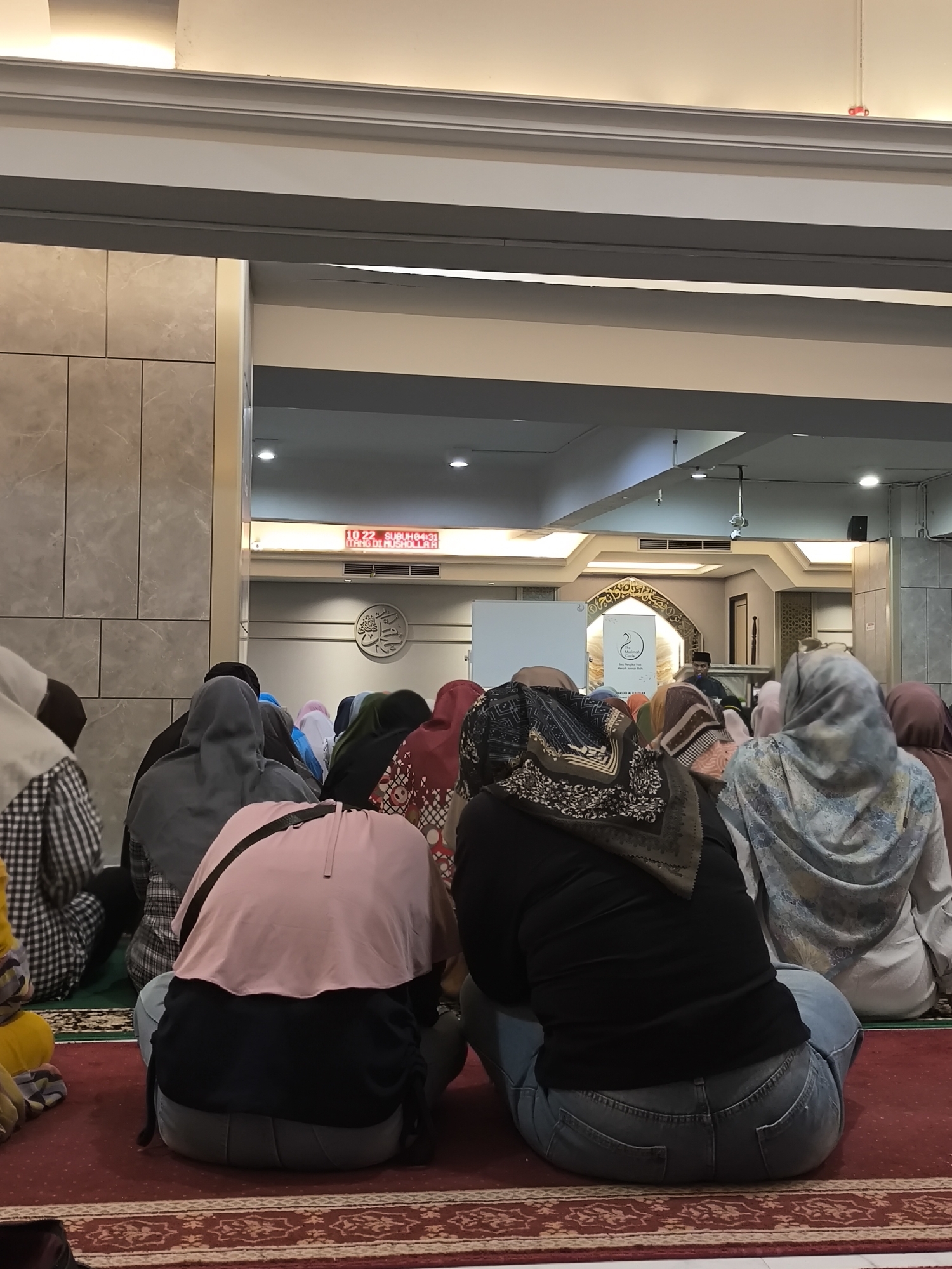 A group of Muslim women, seen from behind, are sitting on a red carpet in prayer at a mosque in Grogol, Indonesia. The women are wearing hijabs and modest clothing. A digital clock on the wall displays "10.22 SUBUL84:31" and "TANG DI MUSHOLL A R". The mosque's interior features tiled walls and a decorative archway with Arabic calligraphy. The lighting is soft and the atmosphere appears peaceful.