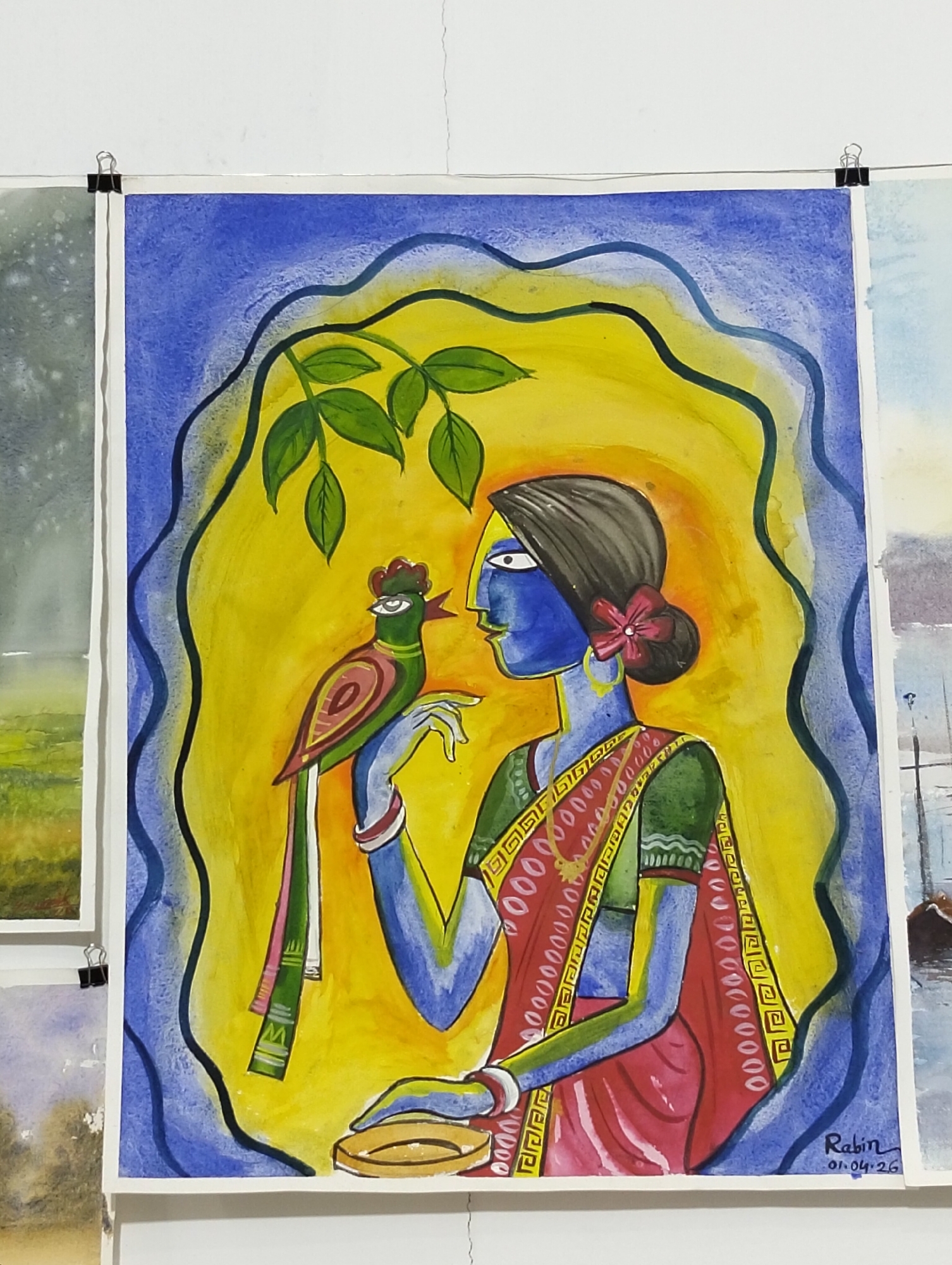 The image shows a painting displayed in a gallery or exhibition space. The painting depicts a woman with blue skin, adorned with a red flower in her dark hair, wearing a green and red sari. She is seated, holding a small, shallow bowl in her left hand. A green and red bird is perched on her right hand. The background is a yellow oval bordered by a blue, wavy line. Above the woman and bird are green leaves on branches. The painting is signed "Rabin 01.04.26" in the lower right corner. The painting appears to be watercolor on paper. The setting of the location being in the city of Dhaka, Bangladesh is not shown or known from the image.