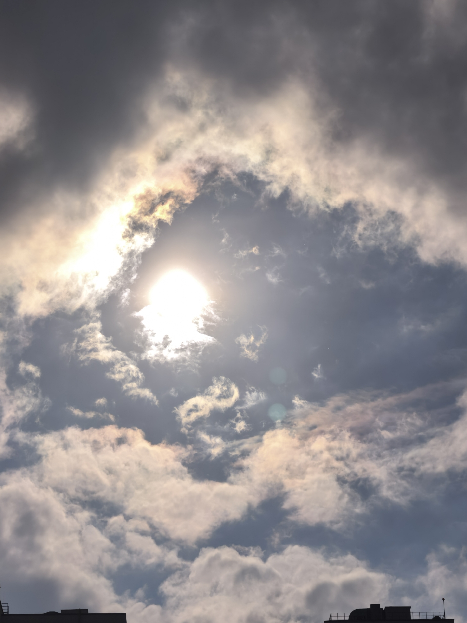 The image captures a dramatic sky over an urban landscape. The sun is partially obscured by thick, swirling clouds, creating a halo effect. The clouds are a mix of dark gray and bright white, with hints of pink and yellow where the sunlight breaks through. The lower portion of the image shows the silhouettes of buildings against the sky, suggesting an urban setting. The composition is dominated by the sky, with the buildings serving as a grounding element. The overall mood is one of awe and a touch of mystery, as the sun battles through the dense cloud cover.