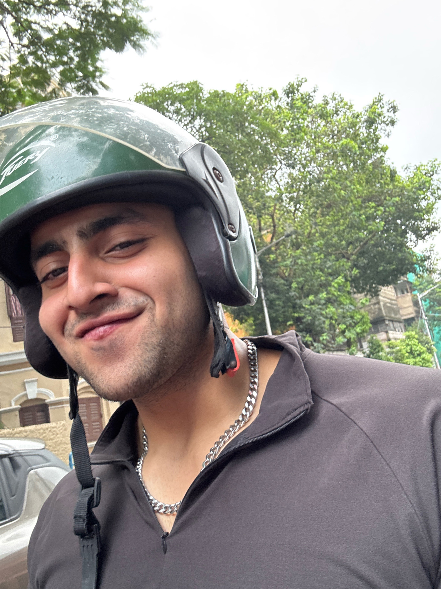 The image shows a close-up, selfie-style shot of a young South Asian man wearing a green helmet and a dark jacket. He is smiling and looking directly at the camera. The man has dark hair and a beard. He is wearing a silver chain necklace. In the background, there are trees, a building with balconies, and a car. The presence of the trees and architectural style of the building suggest an urban environment. While the image itself does not explicitly state the location, the user has specified Kolkata, India.