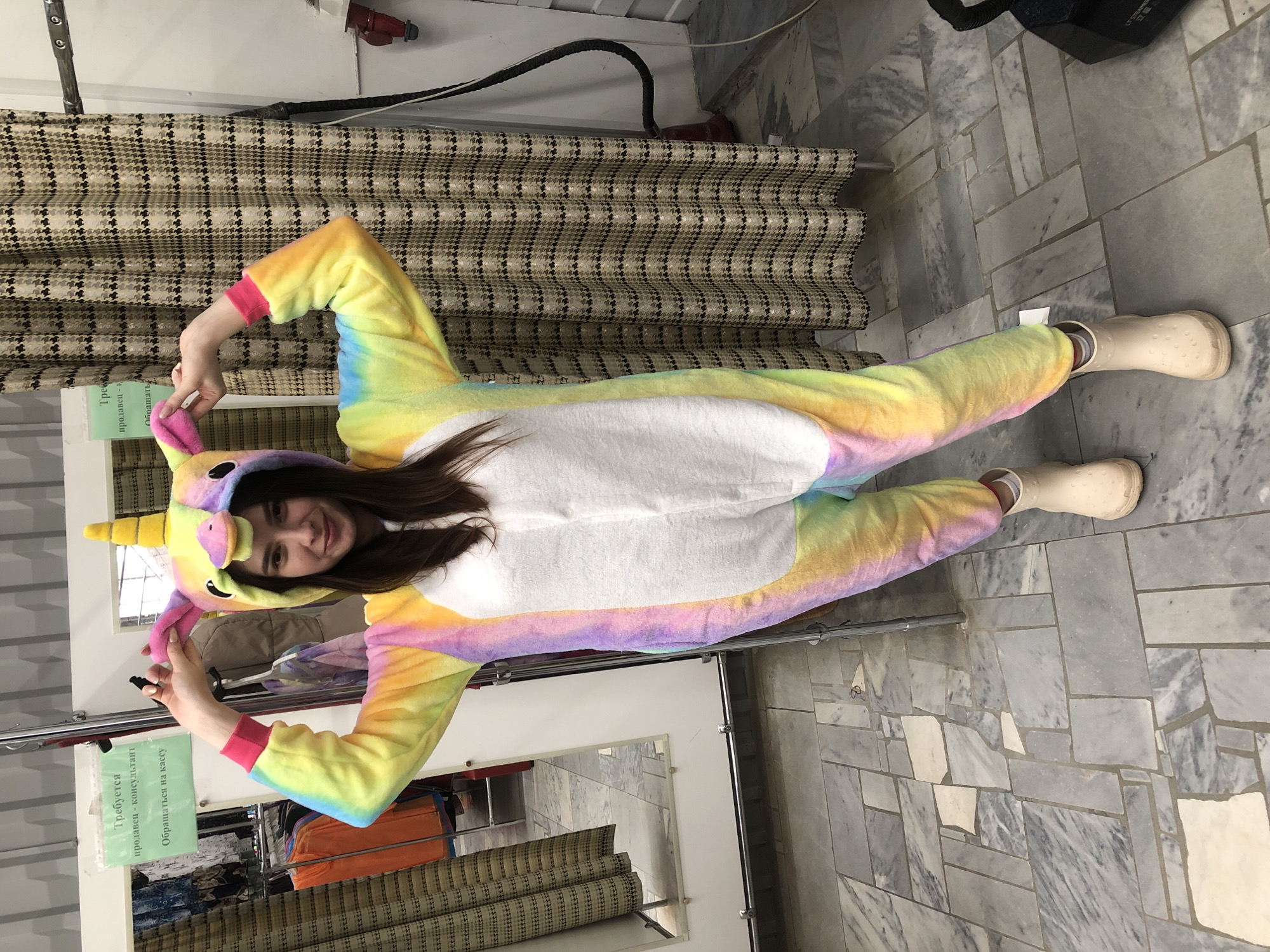 A young woman is lying on her back on a floor covered with grey and white marble tiles, wearing a rainbow unicorn onesie with a white belly and matching beige clogs. She is smiling and making a heart shape with her hands near her head.
The location appears to be a retail store, possibly a clothing store or a fabric store. In the background, there is a clothing rack with clothes and a mirror. A sign in Russian is visible on the rack. Above the rack, there is a patterned fabric. A red electrical outlet and cable are visible at the top of the frame. The city is Chaikovsky, Russia.