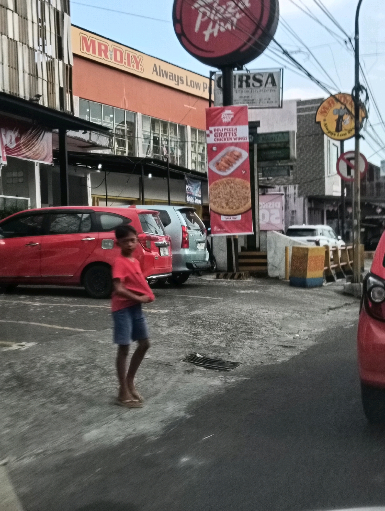 The image captures a street scene in Tomohon, Indonesia, under an overcast sky. In the foreground, a young boy wearing a red t-shirt and blue shorts walks across a wet, paved surface, his feet clad in flip-flops. The pavement appears to be a parking lot, with white markings visible. To the left, a red car is parked, and behind it, another car, possibly silver, is also present. A red banner displaying images of pizza and chicken wings, along with text in what appears to be Indonesian, is prominently displayed on a pole. Above the banner, signs for "MR. DIY" and "Pizza Hut" are visible on a building with an orange facade. Further in the background, more buildings and electrical poles with tangled wires contribute to the urban landscape. The overall impression is of a commercial area on a day with possibly recent rainfall.