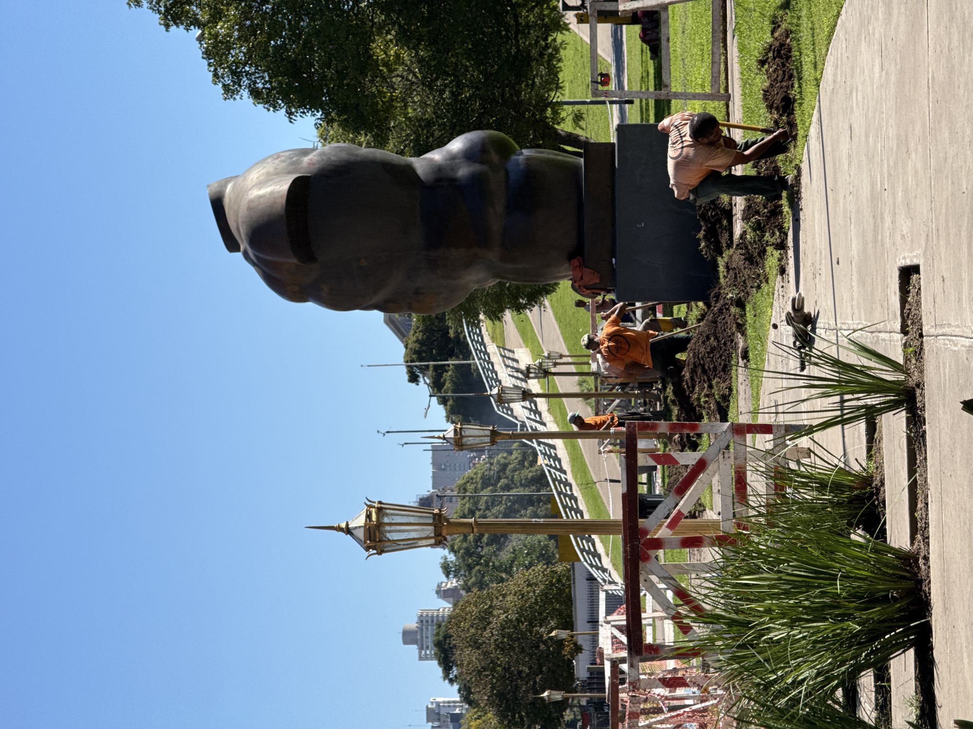The image depicts an outdoor scene in what appears to be a public park or plaza in Buenos Aires, Argentina, under bright, clear daylight. The main subject is a large, dark, abstract or highly stylized sculpture, resembling a human head or torso, mounted on a dark, rectangular pedestal.
Around the base of this sculpture and along a paved pathway to its right, several workers are engaged in landscaping or maintenance activities. One worker, wearing an orange or light brown t-shirt and dark trousers, is prominently shown actively digging with a shovel, creating piles of dark soil along the edge of the concrete path. Another worker in a similar orange shirt is visible further back, near the sculpture's base, along with a partially obscured wheelbarrow. Red and white striped wooden barriers are set up along the path, indicating a work zone.
The scene includes lush green grass, several mature trees, and elegant, ornate street lamps with golden tops, which line another pathway in the background. Beyond these elements, distant urban buildings are visible against a clear blue sky, suggesting an urban park setting. The overall activity conveys ongoing public infrastructure maintenance or aesthetic improvements. The time of day is sunny mid-day, and the weather is clear. No discernible text is clearly visible on any signs or clothing.