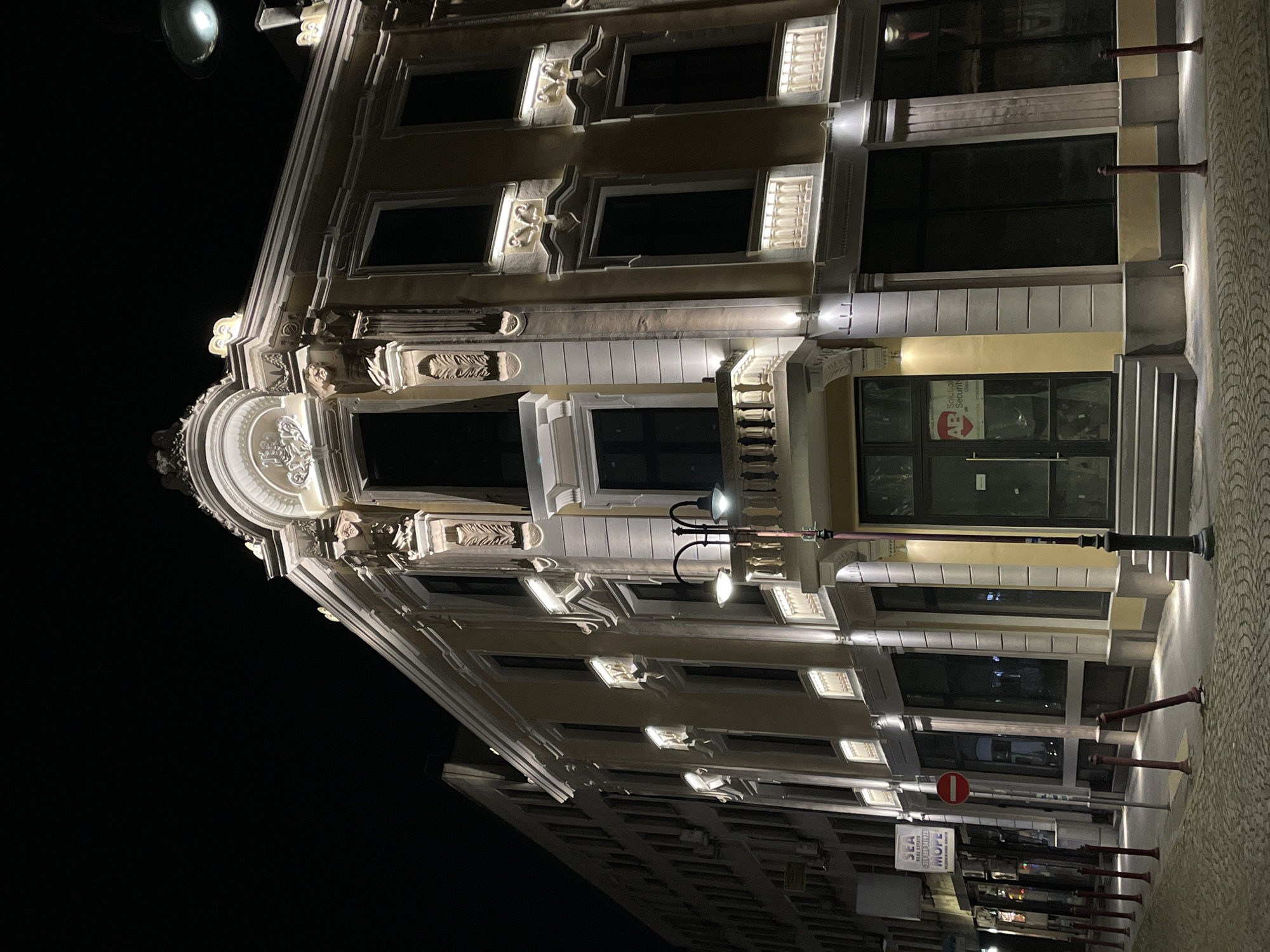 A low-angle nighttime shot features a well-lit ornate building in Burgas, Bulgaria. The building's facade has decorative elements around the windows and along the roofline. The windows are dark, suggesting that the building is not currently occupied. On the ground floor, there is an entrance with glass doors. A street lamp stands near the entrance. Sidewalk with street posts, a sign, and other urban details are also visible.